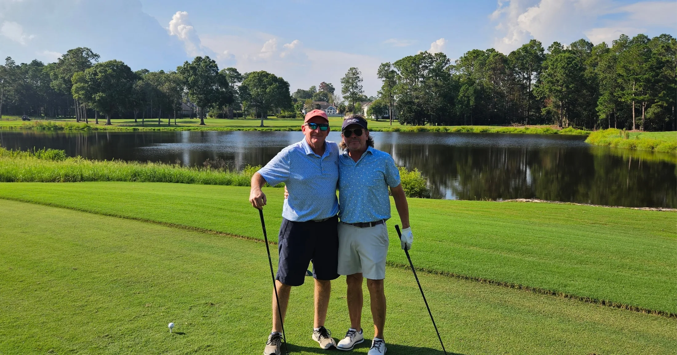 Chuck Kelly and David Boggs posing together with clubs on the fairway, a pond reflecting the treeline behind them