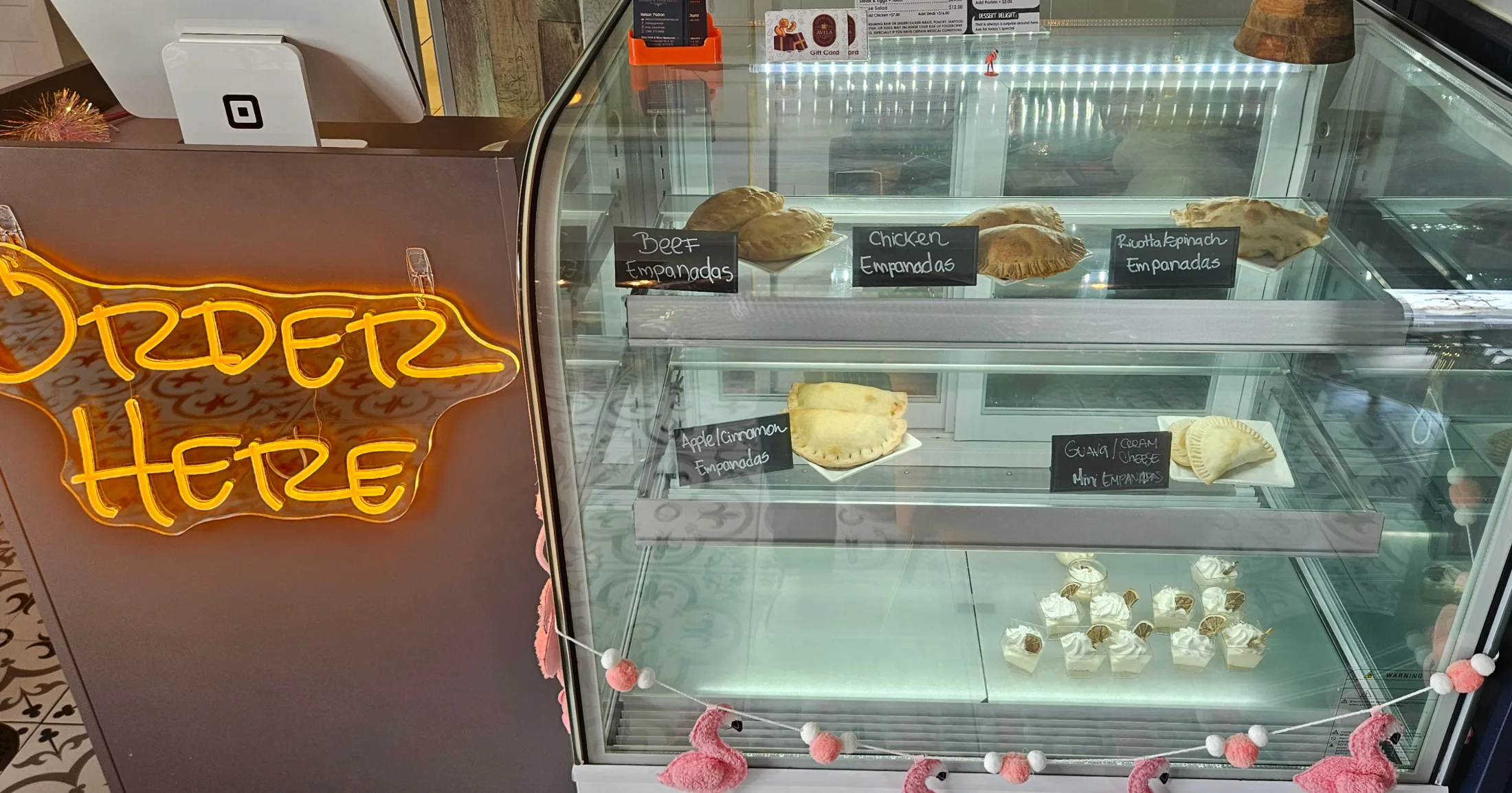 Glass display case at Avila showing beef, chicken, ricotta spinach, apple cinnamon, and guava cream cheese empanadas alongside small desserts