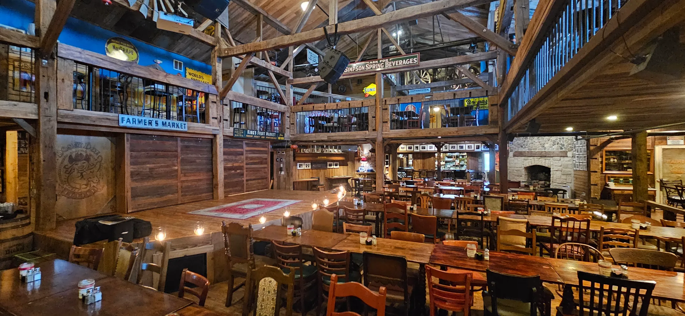Wide interior view of Dosey Doe showing the full barn space, vintage signage including Farmer's Market and King's Fruit Market, stone fireplace, and the lit performance stage