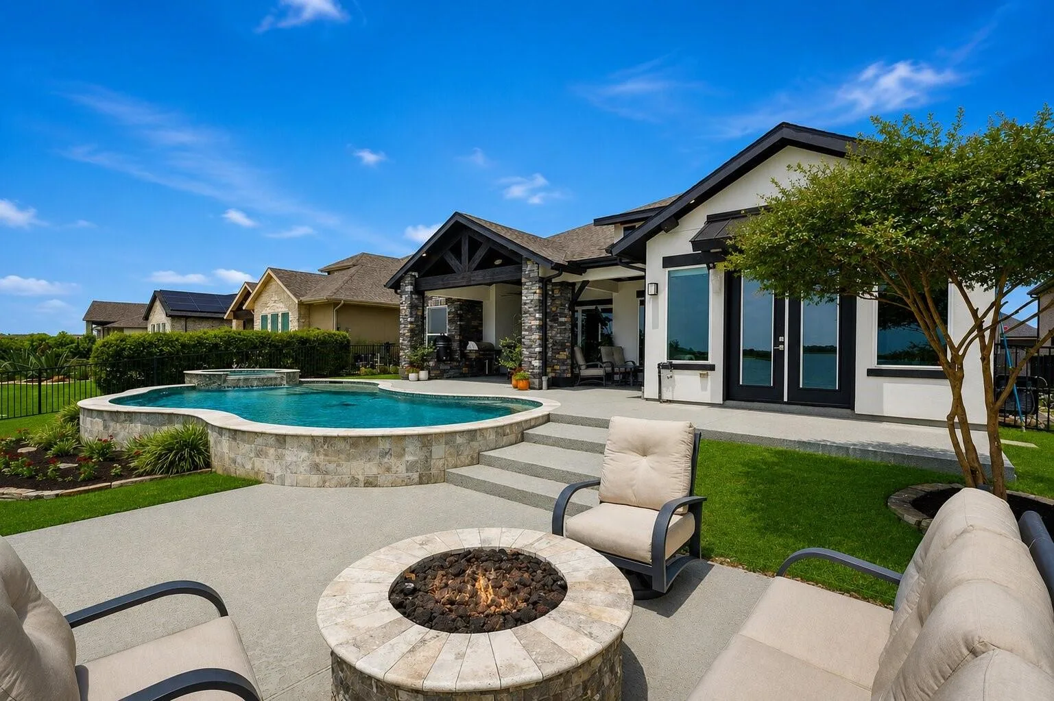 Rear exterior showing covered patio with stone columns, fire pit seating area, elevated pool deck, and lake in the background