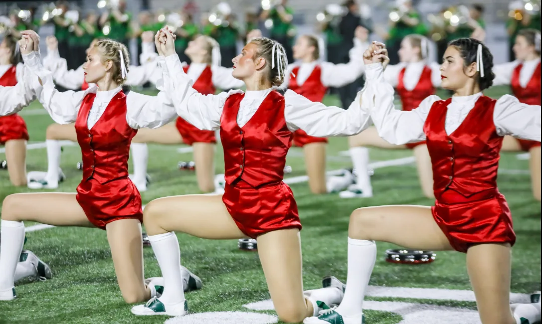 TWHS Highsteppers perform in red and white costumes on the field at night, kneeling in formation with the marching band behind them