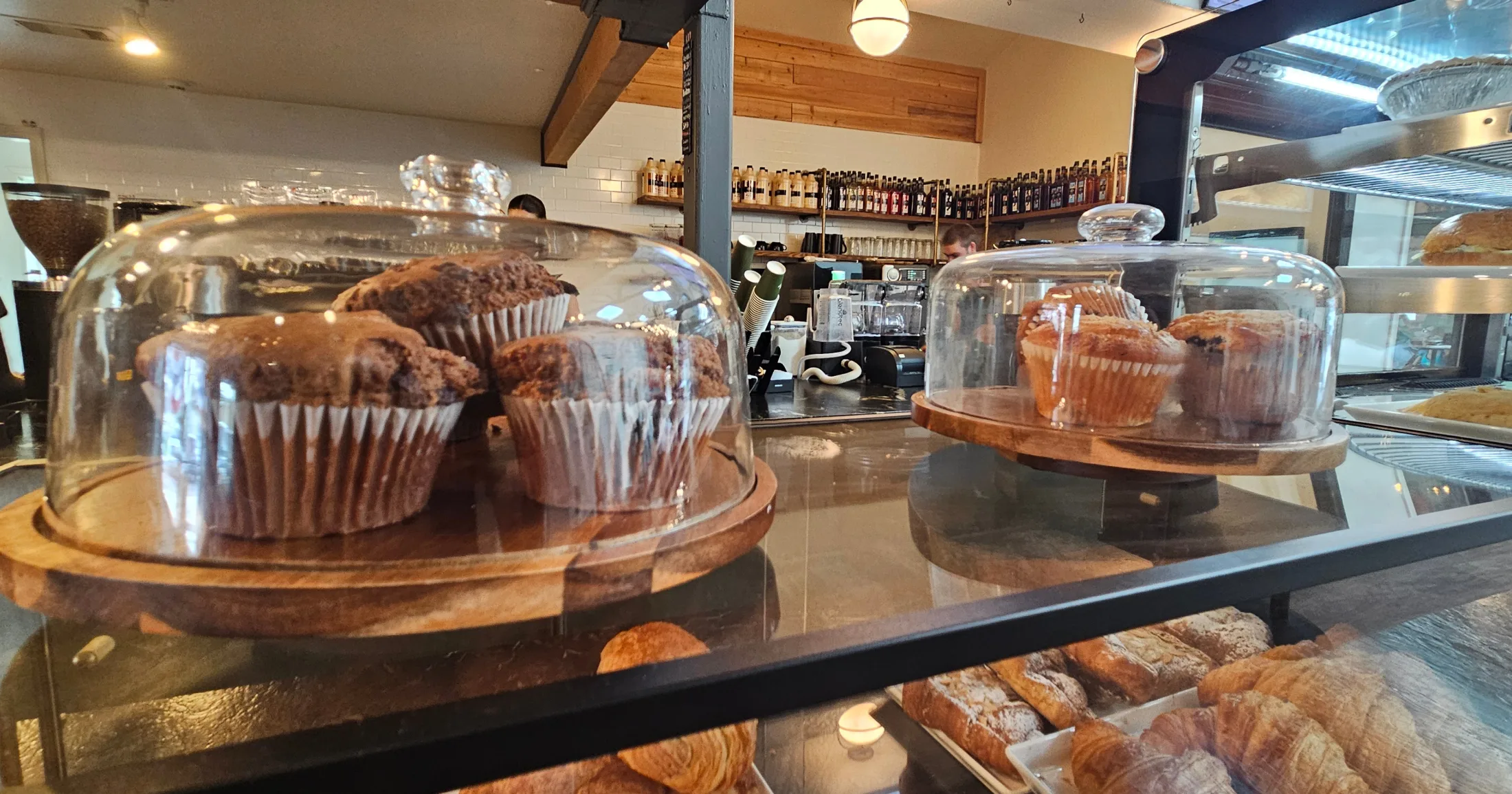 Close-up of muffins under glass domes at First Cup Coffee Co. counter, with croissants visible in the case below