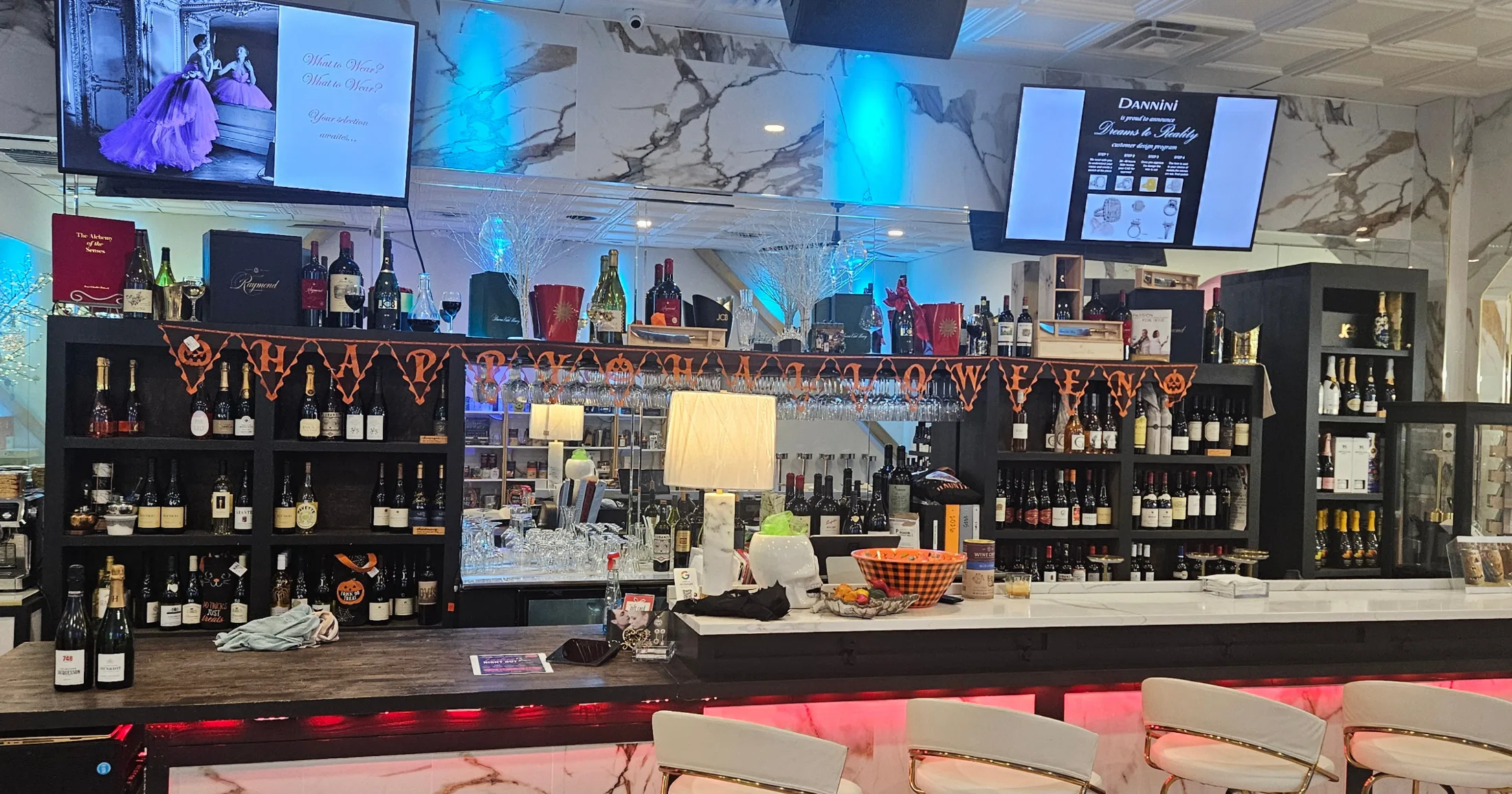 Dannini bar wall stocked with wine bottles and glassware, Halloween banner stretched across the front, screens above showing the Dreams to Reality custom design program