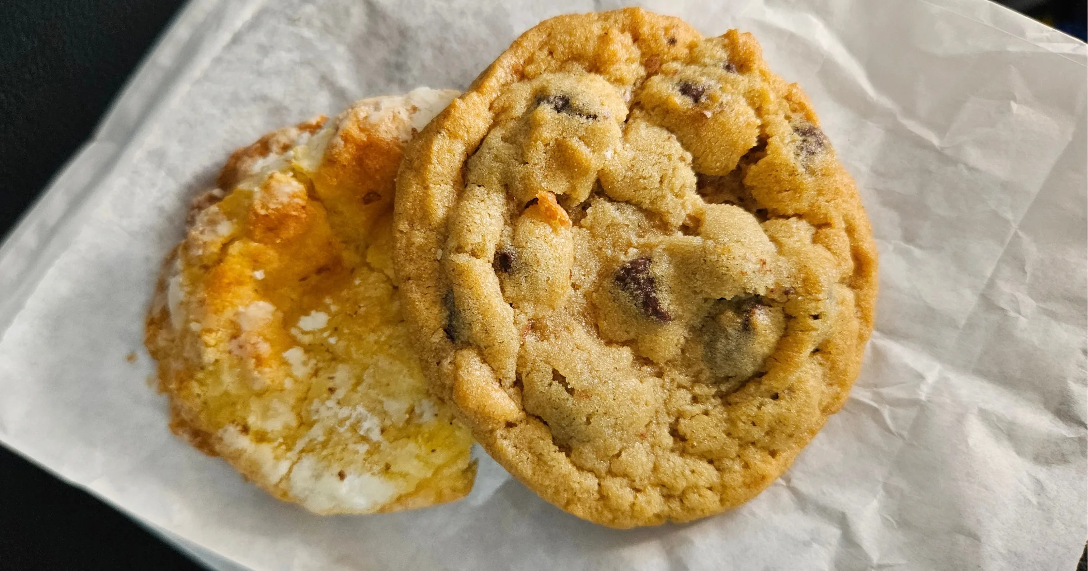 Two freshly baked cookies on parchment paper at The Baked Bear, including a chocolate chip and a sugar cookie style