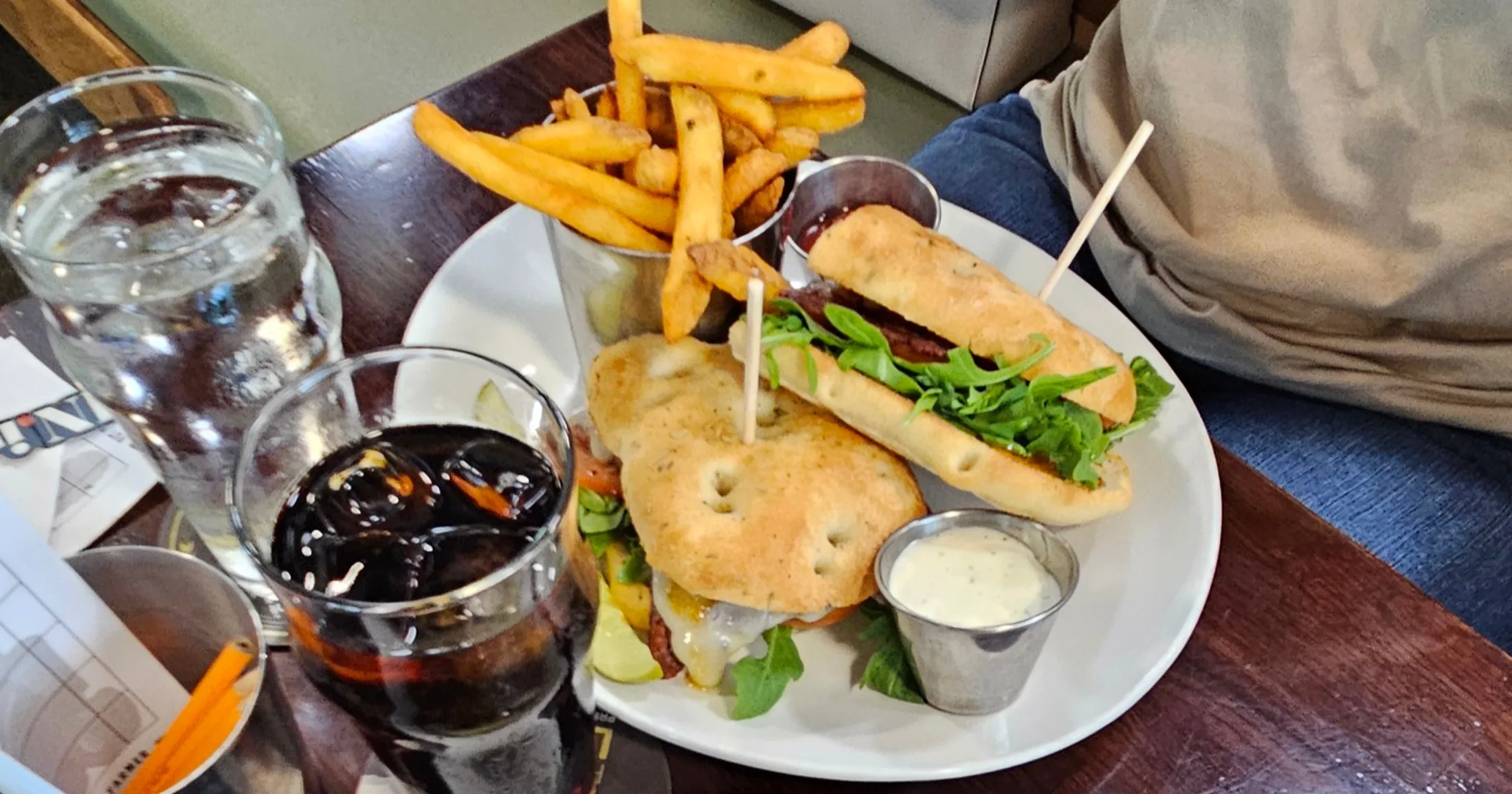 Two sandwiches on focaccia with arugula and melted cheese, fries in a metal cup, and a cold drink at Farmers Brewing Co.