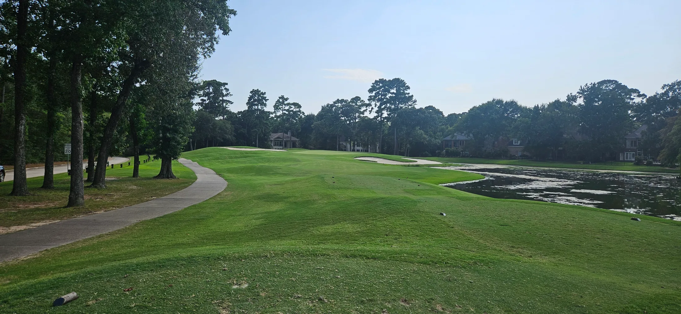 Golf course fairway winding between tall pines with a water hazard on the right, sand traps guarding a green, estate homes visible through the tree line