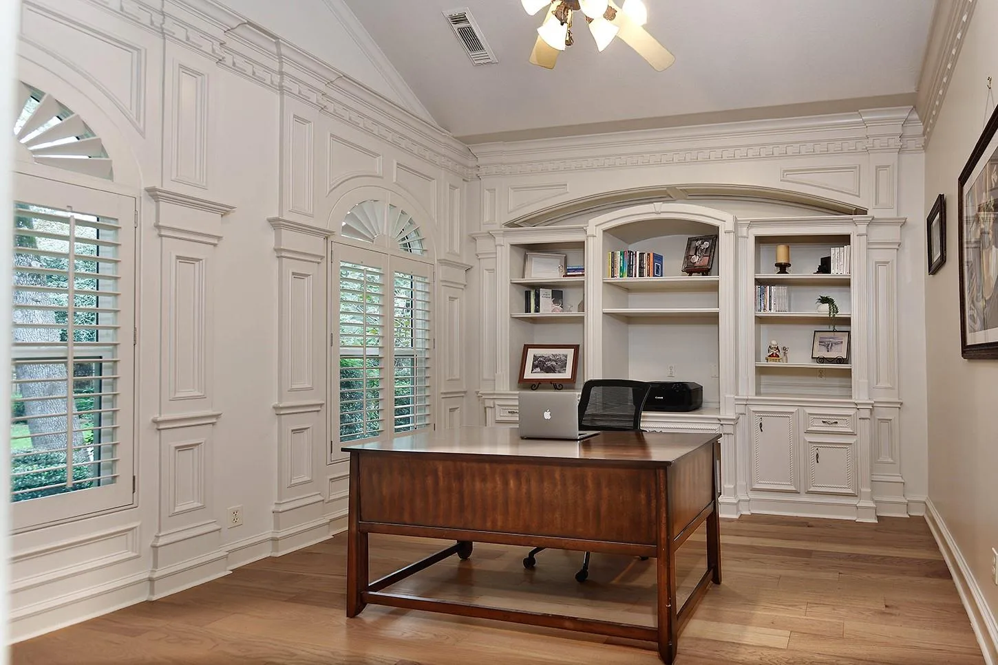 Private study with white wainscoting, built-in bookshelves under arched millwork, plantation shutters, and hardwood floors