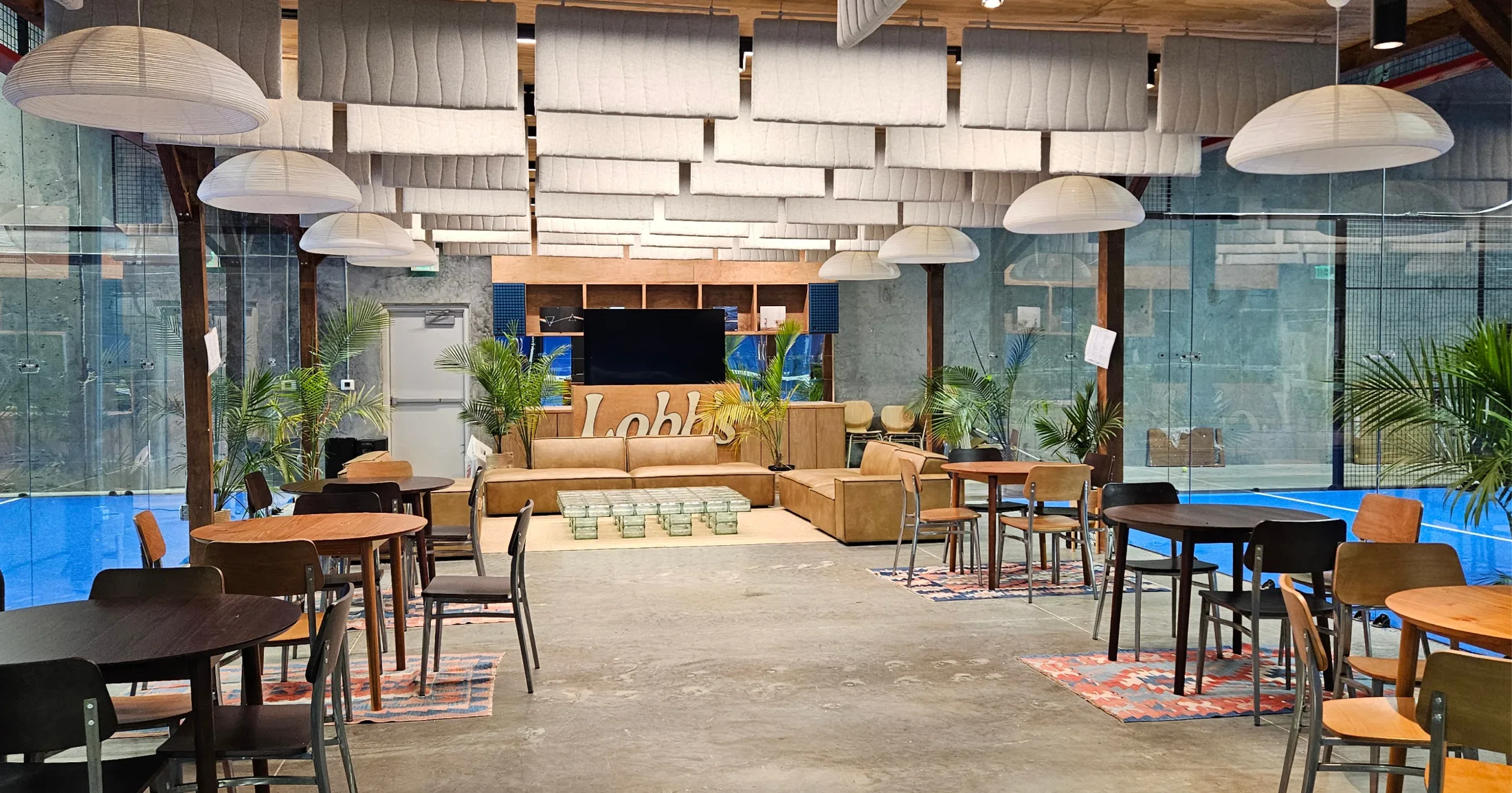 Wide view from inside a padel court looking toward the lounge, with the Wilson net in the foreground, the blue court surface, and the café seating and pendant lights visible through the glass behind