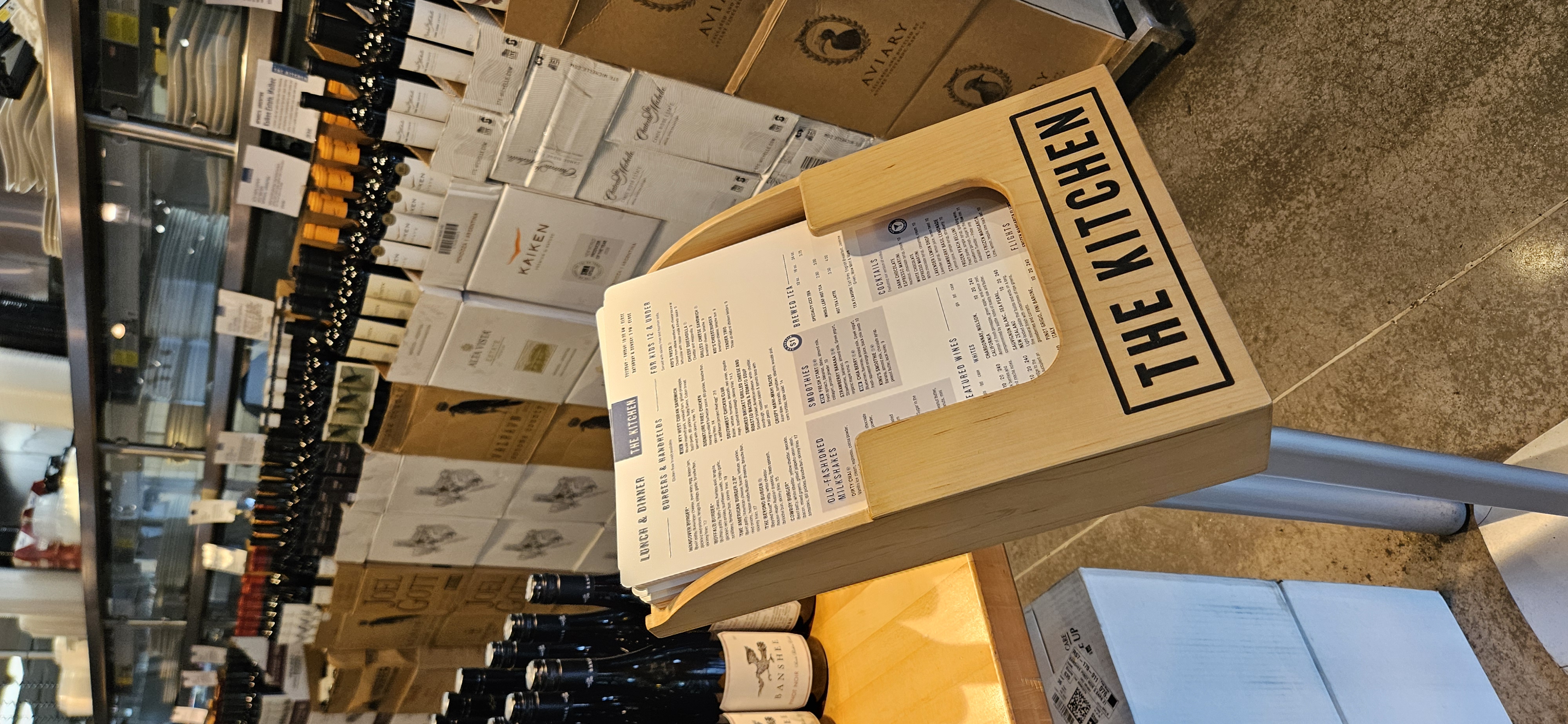 The Kitchen lunch and dinner menu displayed on a wooden stand surrounded by stacked wine bottles and cases