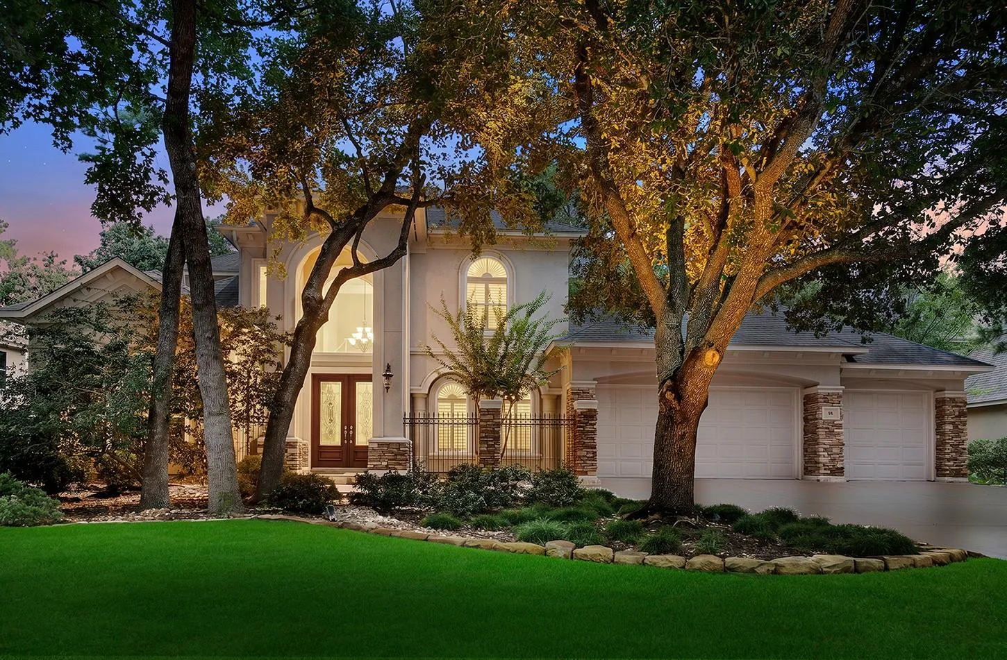 Twilight exterior of 10 Snow Pond Place showing stucco-and-stone facade, arched entry, and mature oak canopy