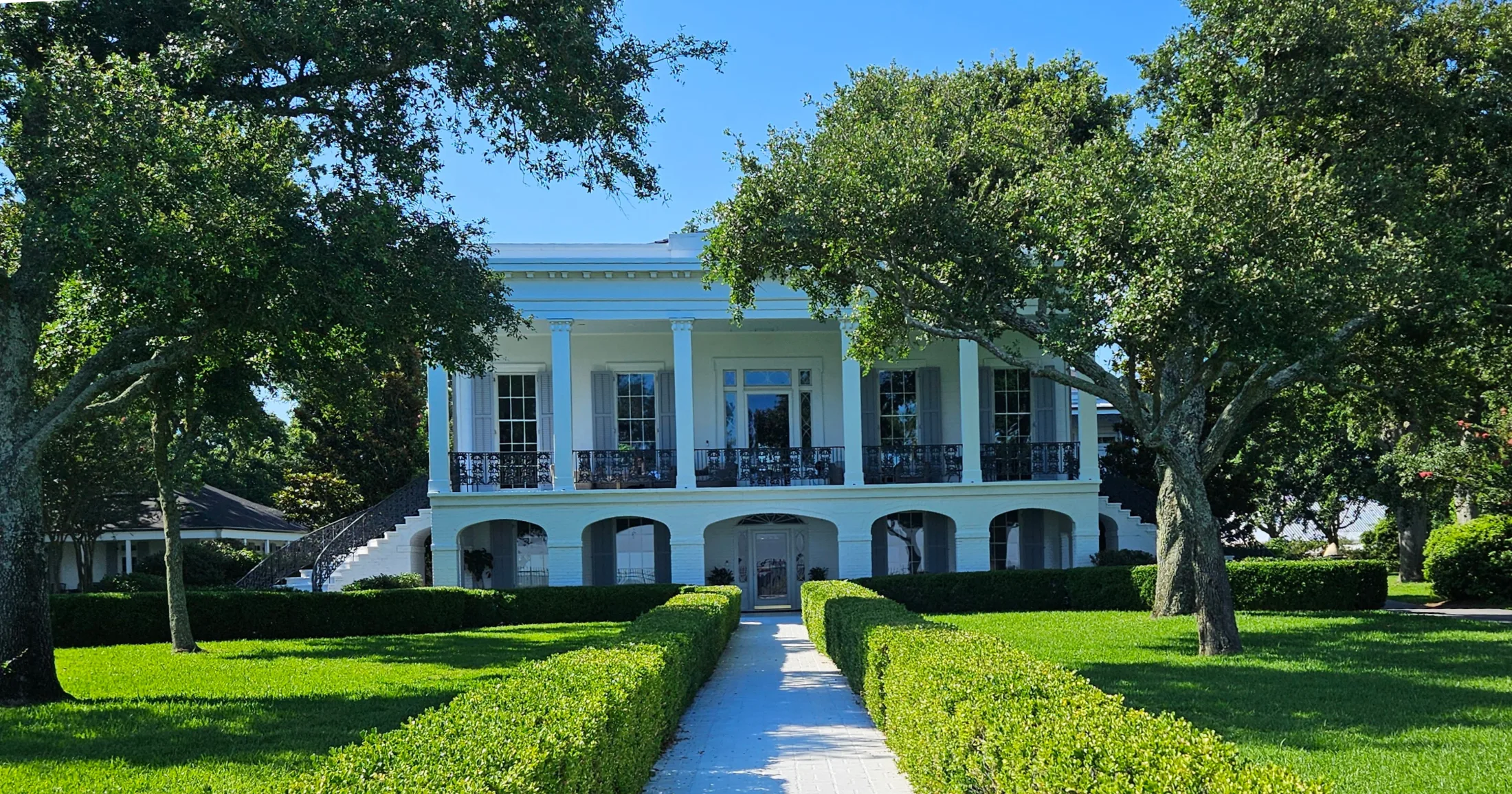A formal white antebellum-style home with arched lower colonnades, ornate iron railings on the upper balcony, and a manicured boxwood hedge pathway