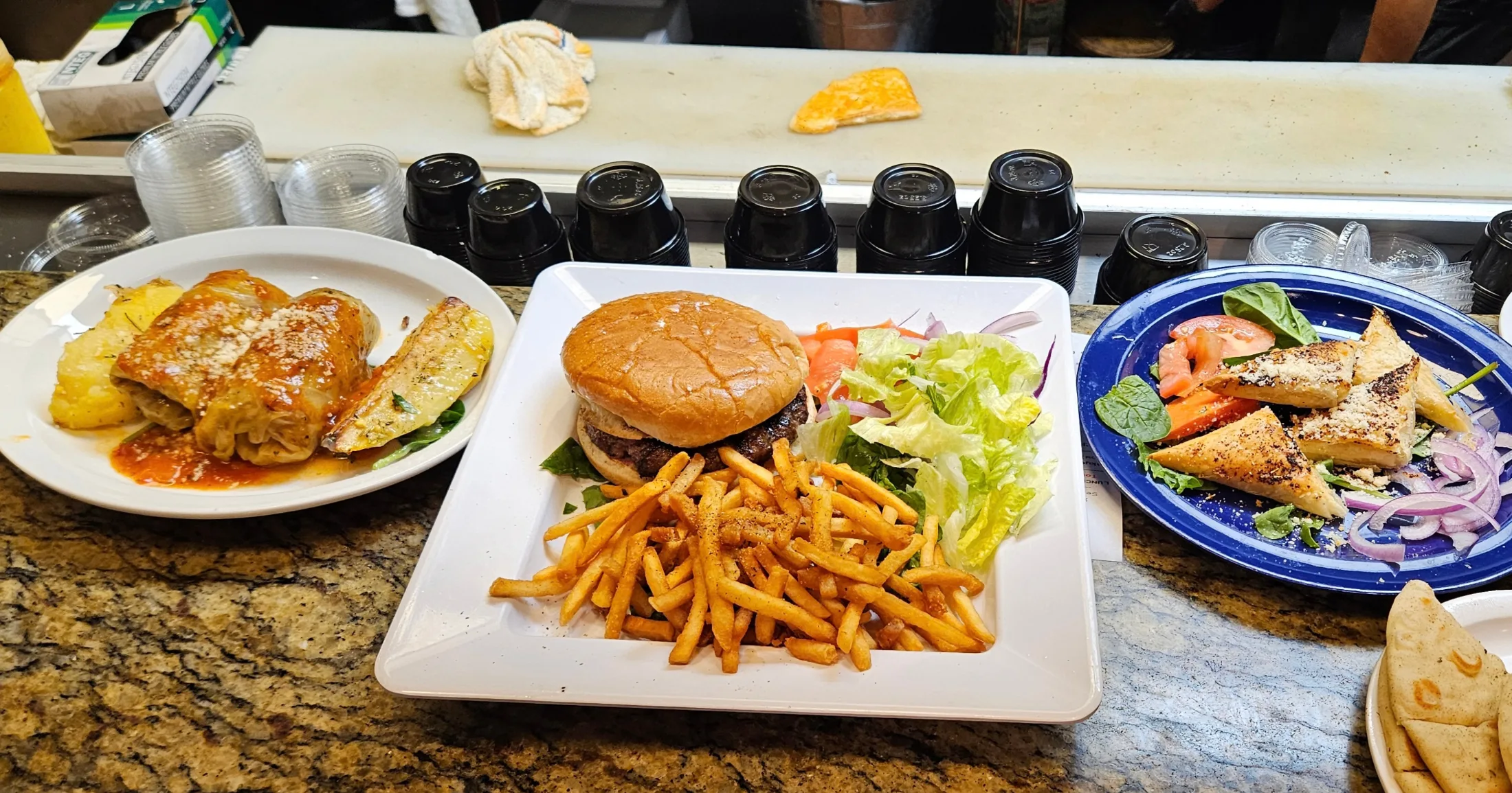 Counter view of multiple plates including pastitsio, a burger with fries, and a Greek salad with pita