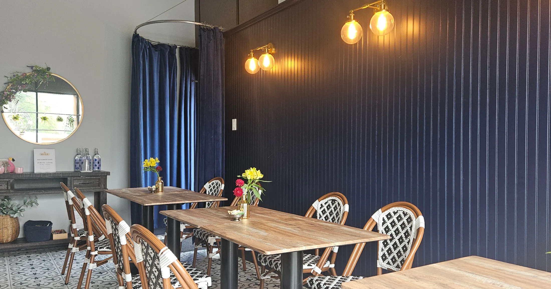Avila Coffee Shop dining room with navy paneled wall, warm globe sconces, wood tables and woven bistro chairs
