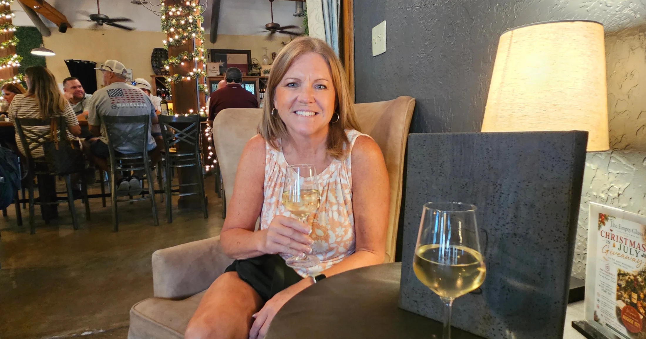 Woman relaxing in a lounge chair at The Empty Glass with two glasses of white wine, full dining room visible behind her