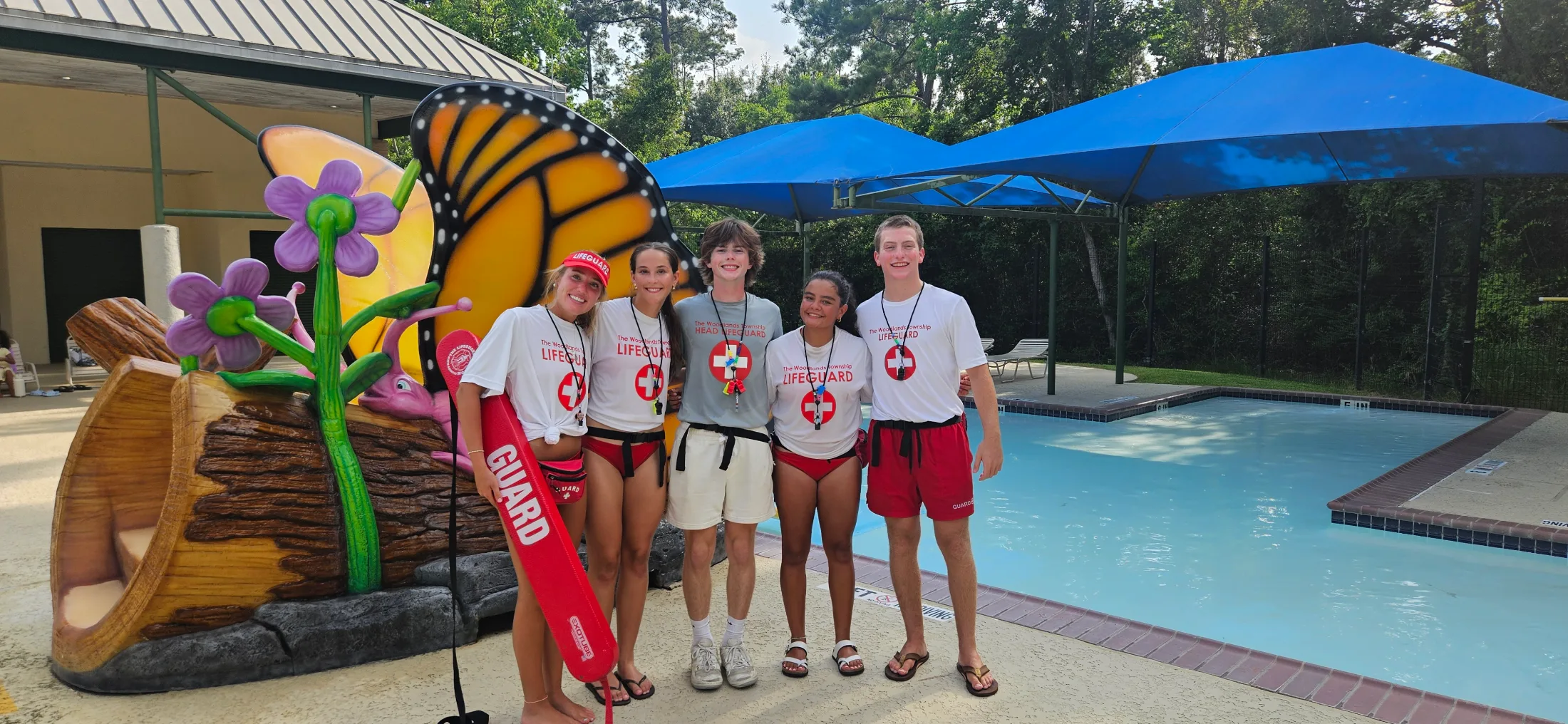 Five Woodlands Township lifeguards posing at a community pool beside a monarch butterfly play structure, one holding a red rescue tube