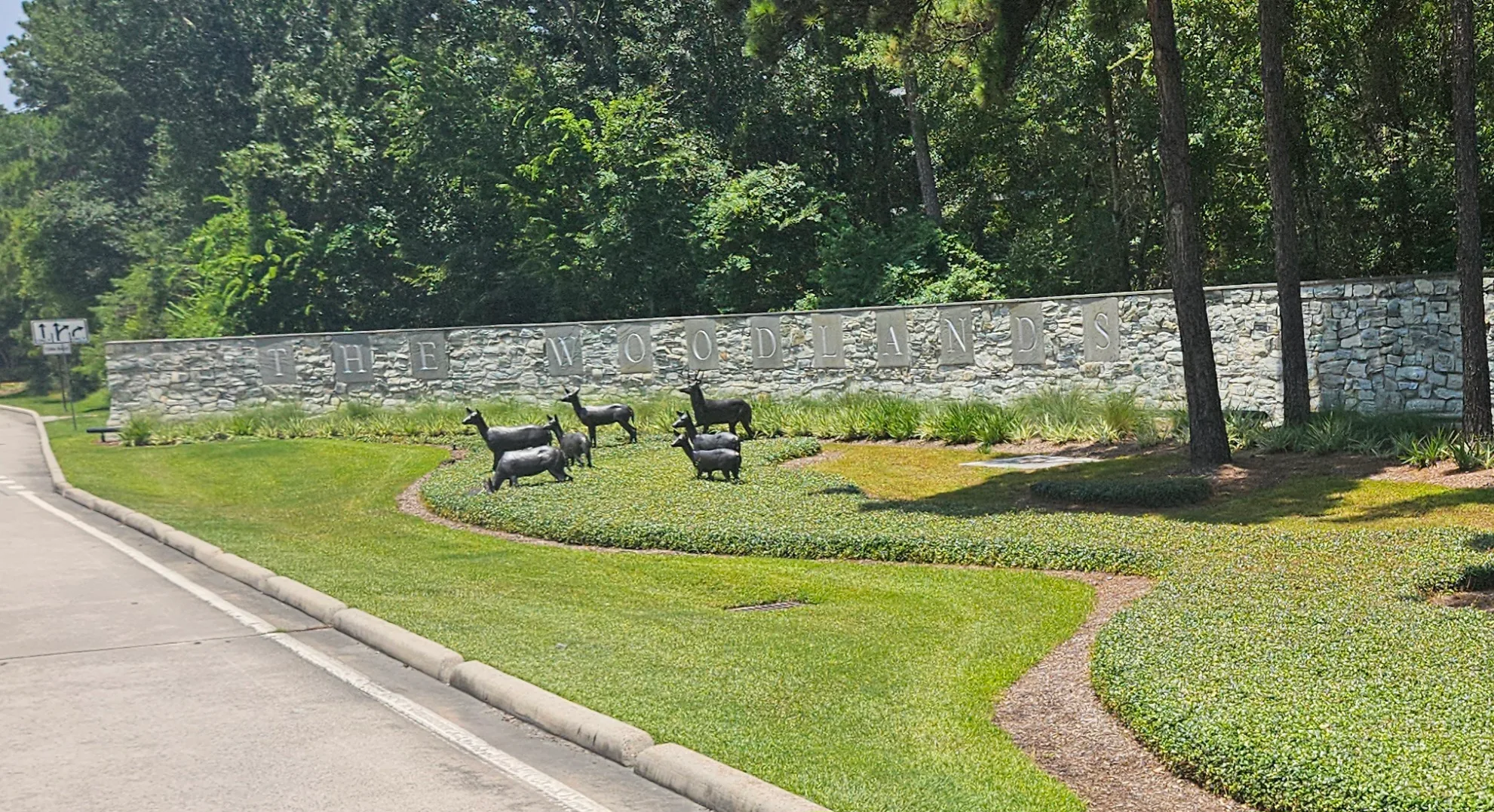 The Woodlands stone entrance wall with bronze deer sculptures mid-stride across the landscaped median, loblolly pines behind