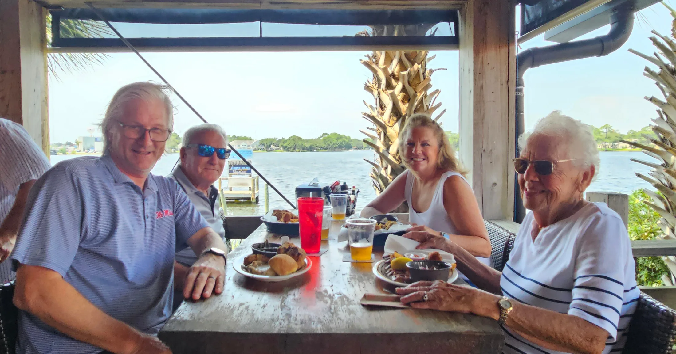 Group of four dining on a covered waterfront patio, cold drinks on the table and the bay behind them