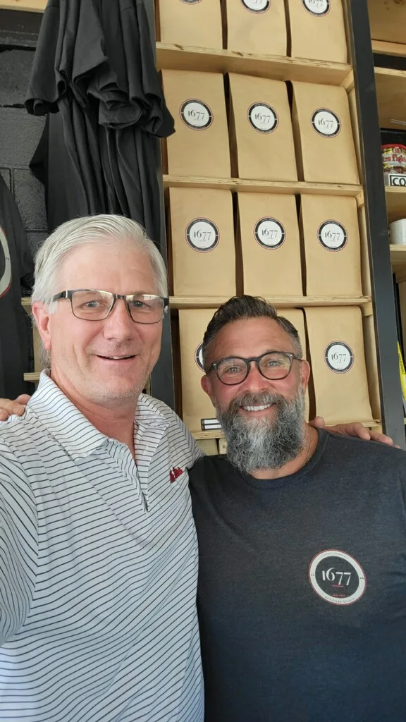 Rick Raanes and David Dunlap standing together in front of a wall of 1677-branded coffee bags