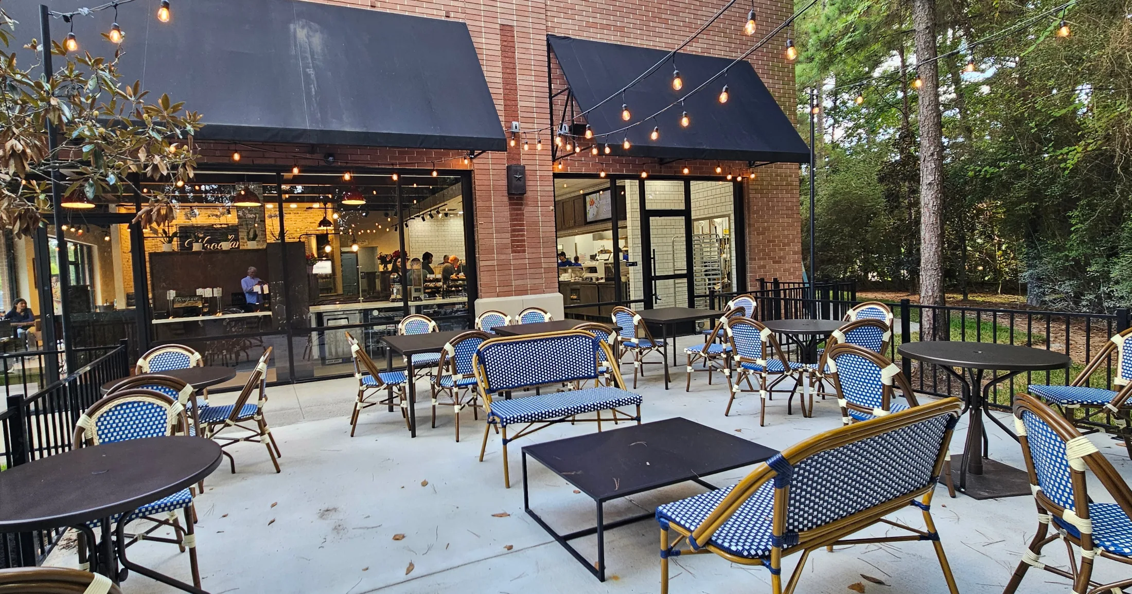 La Madeleine's patio in Alden Bridge at dusk, with blue bistro chairs, string lights, and tall pines in the background