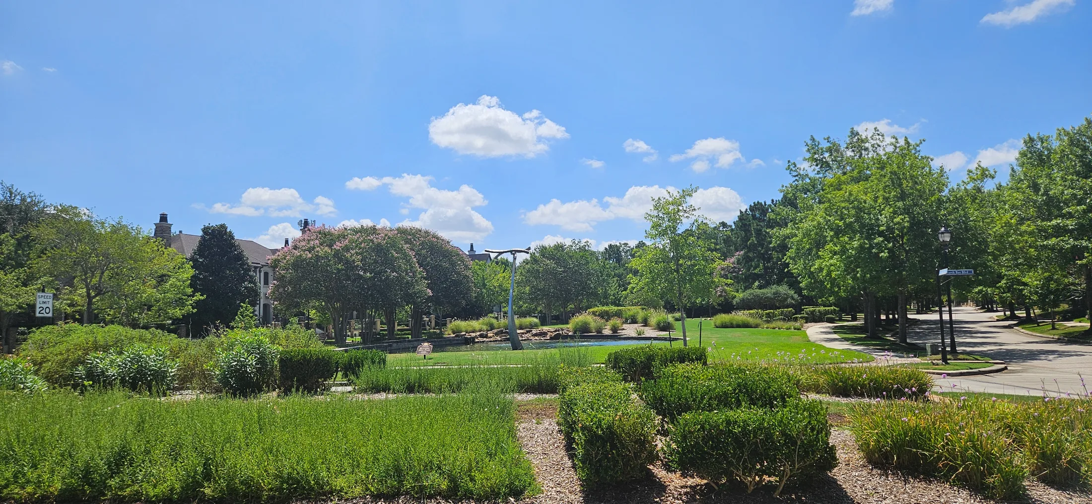 East Shore village green with formal hedgerow garden, reflecting pond, sculpture, and mature trees under a blue Texas sky