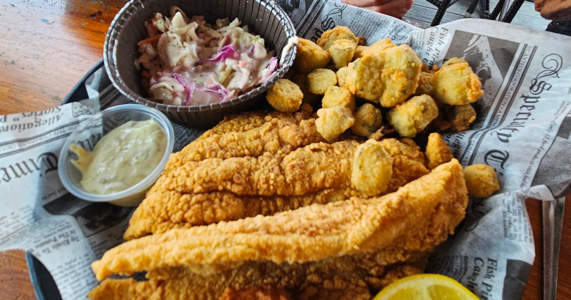 Fried catfish fillets with fried okra, coleslaw, and dipping sauce served on newspaper-lined tray