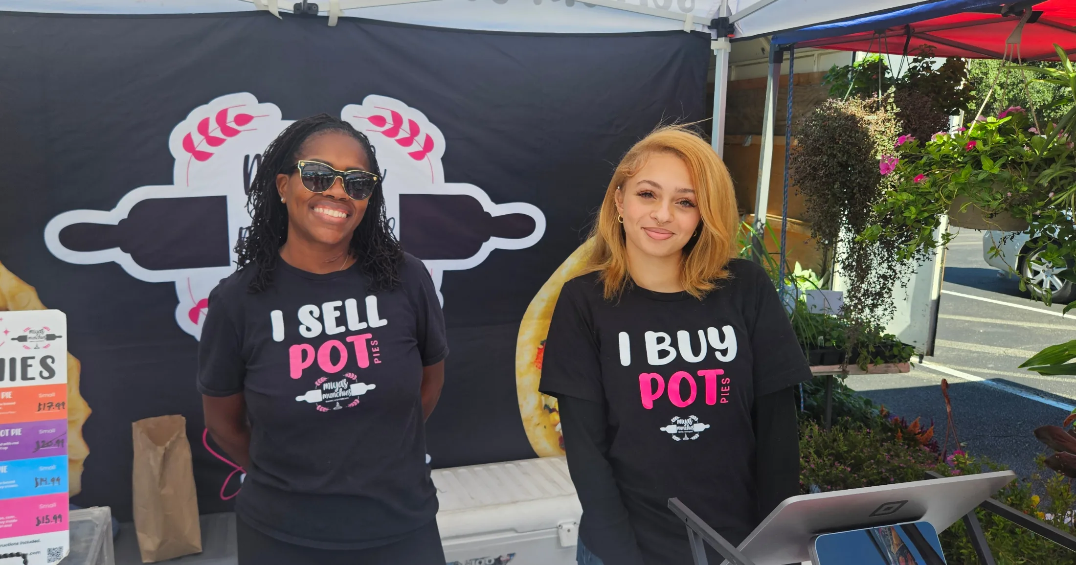 Two women wearing Mya's Munchies shirts reading I Sell Pot Pies and I Buy Pot Pies at a farmers market booth