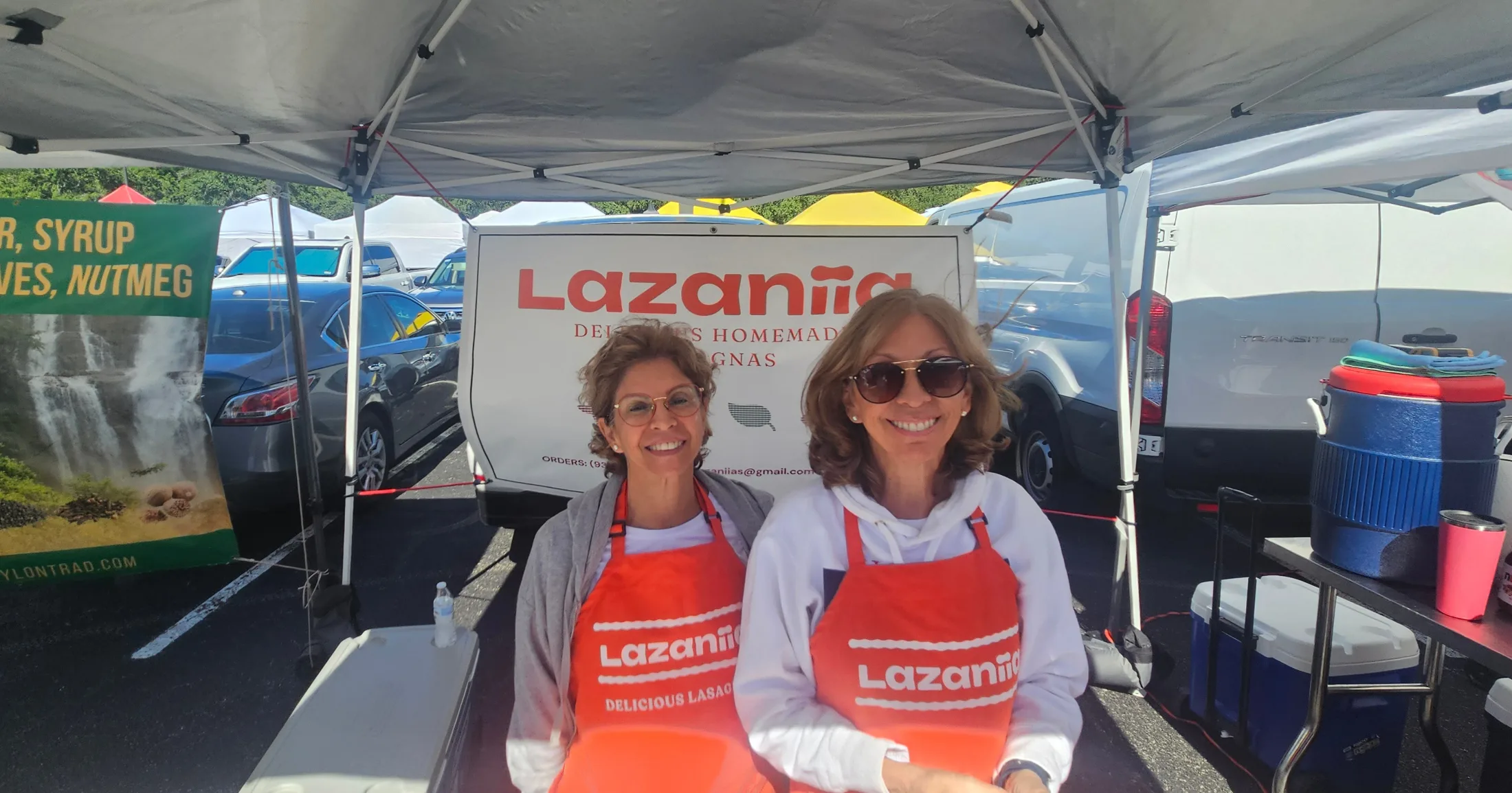 Two women in Lazaniia aprons smiling at their market booth selling homemade lasagnas
