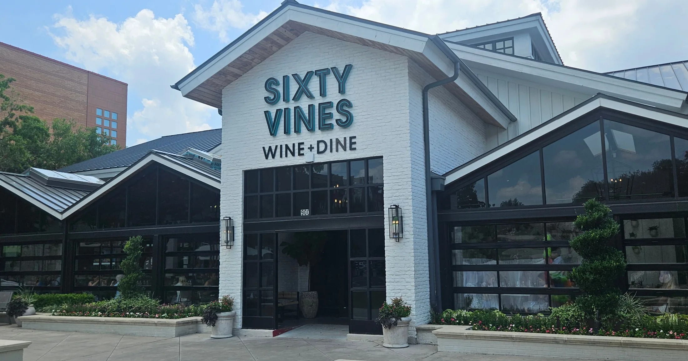 Sixty Vines Wine and Dine restaurant exterior with white brick facade, black metal and glass garage doors, and lush landscaping