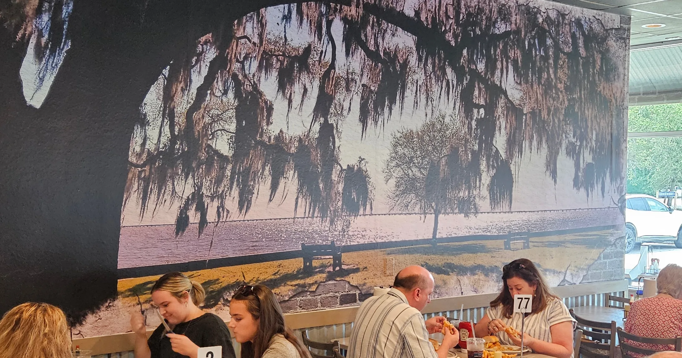Diners eating at tables beneath a large mural of live oaks with Spanish moss overlooking Lake Pontchartrain