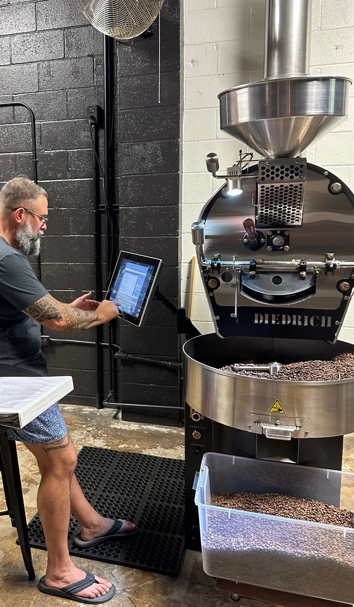 David Dunlap operating a Diedrich commercial coffee roaster, monitoring a roast on a tablet with freshly roasted beans cooling in the drum