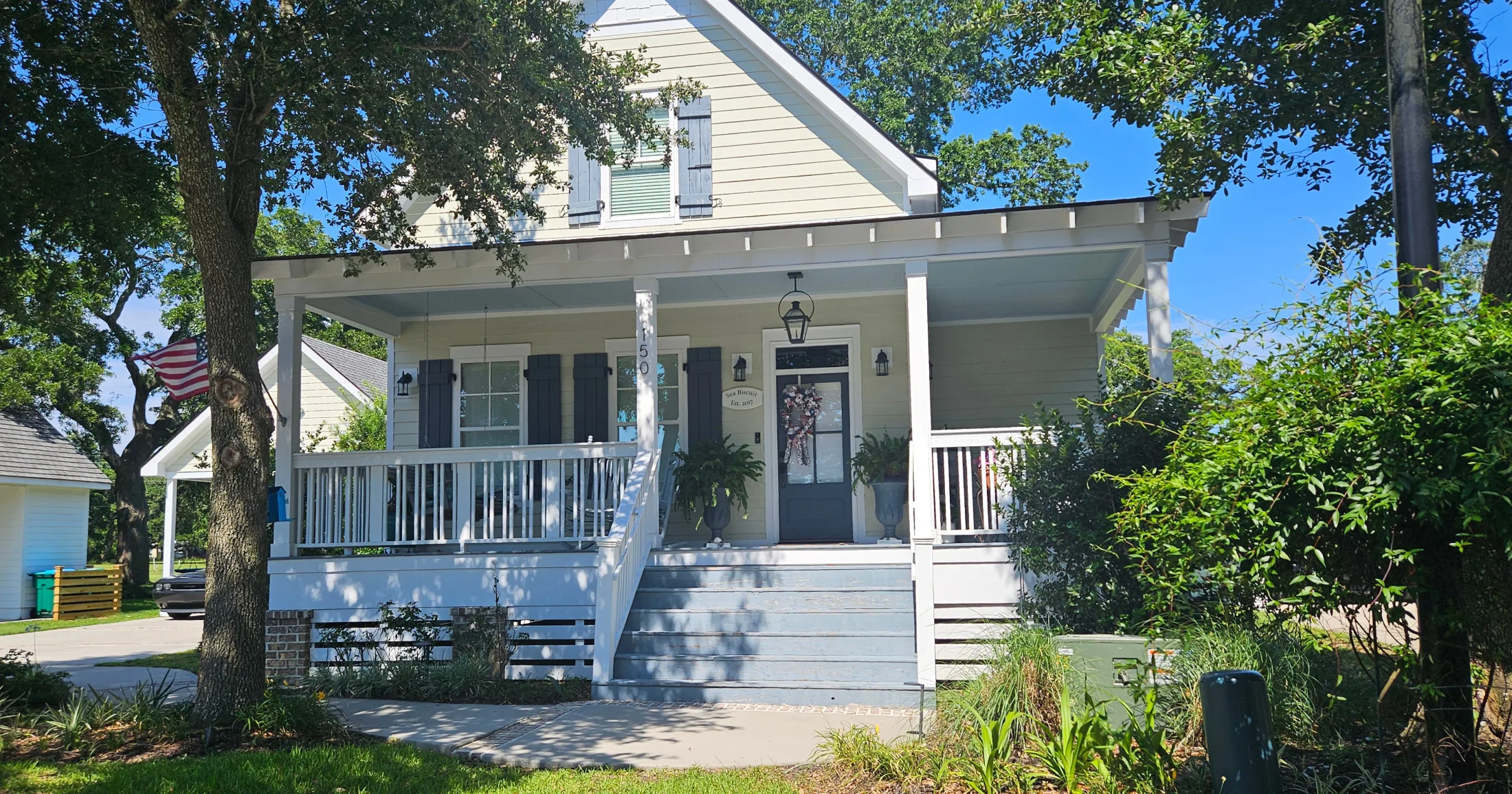 A modest raised coastal cottage painted light blue with a covered front porch, gas lantern, dark shutters, and an American flag β new construction that fits the neighborhood scale