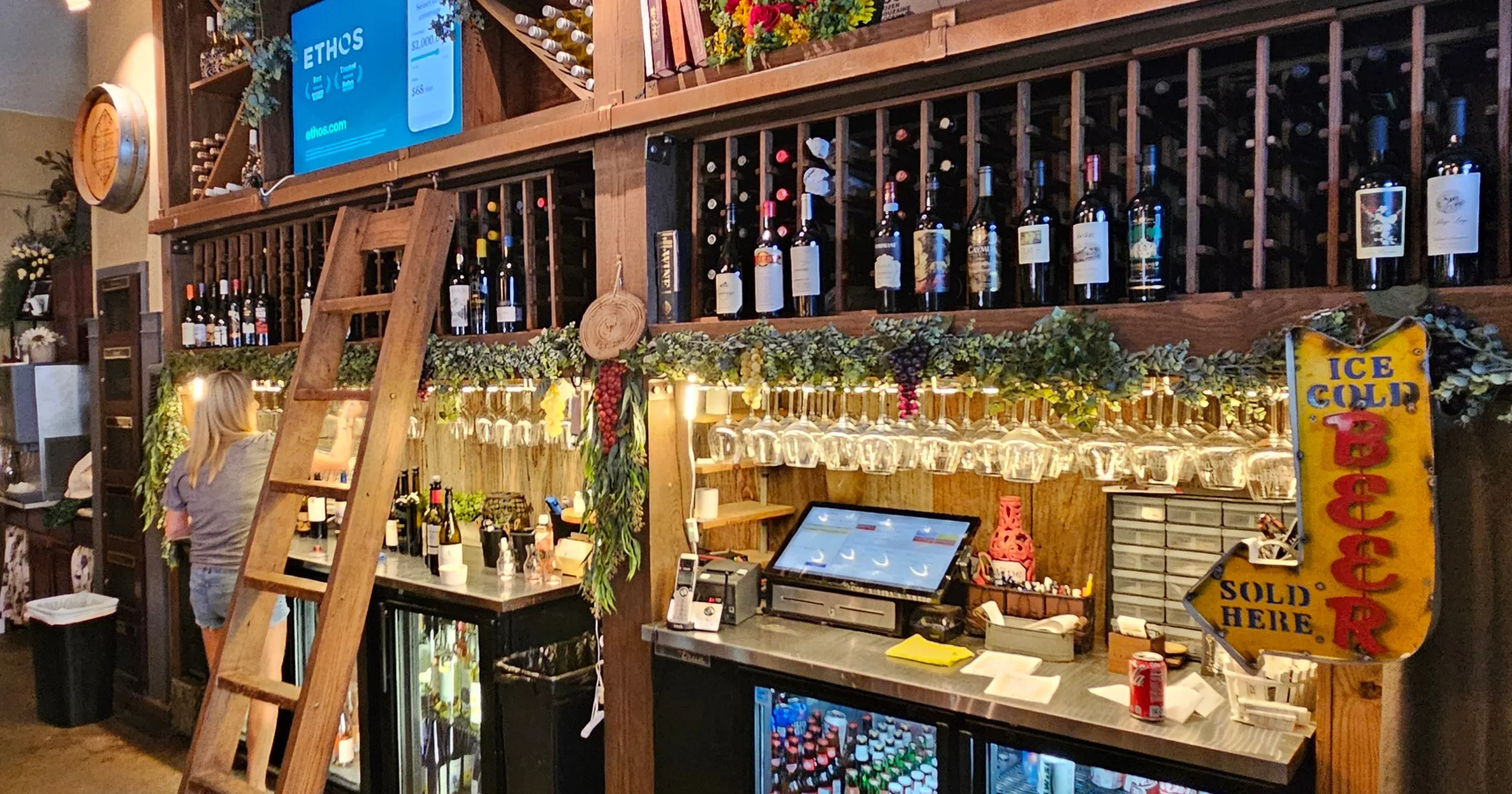Interior bar at The Empty Glass with floor-to-ceiling wine racks, hanging stemware, and a vintage Ice Cold Beer sign