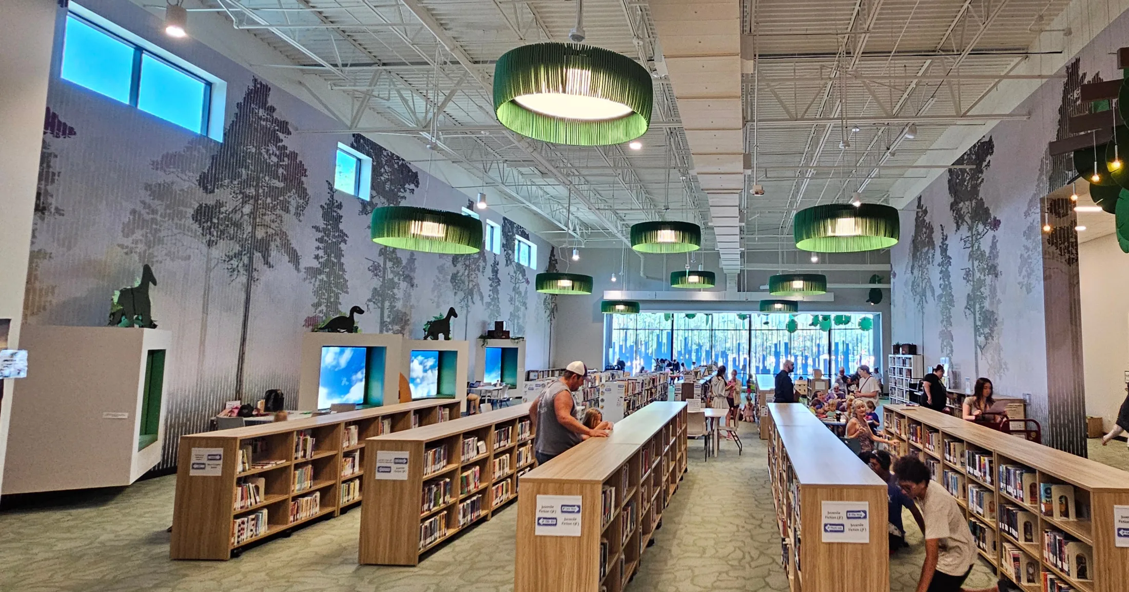 Wide interior view of the Kevin Brady Library showing juvenile stacks, tree-themed murals, and families browsing
