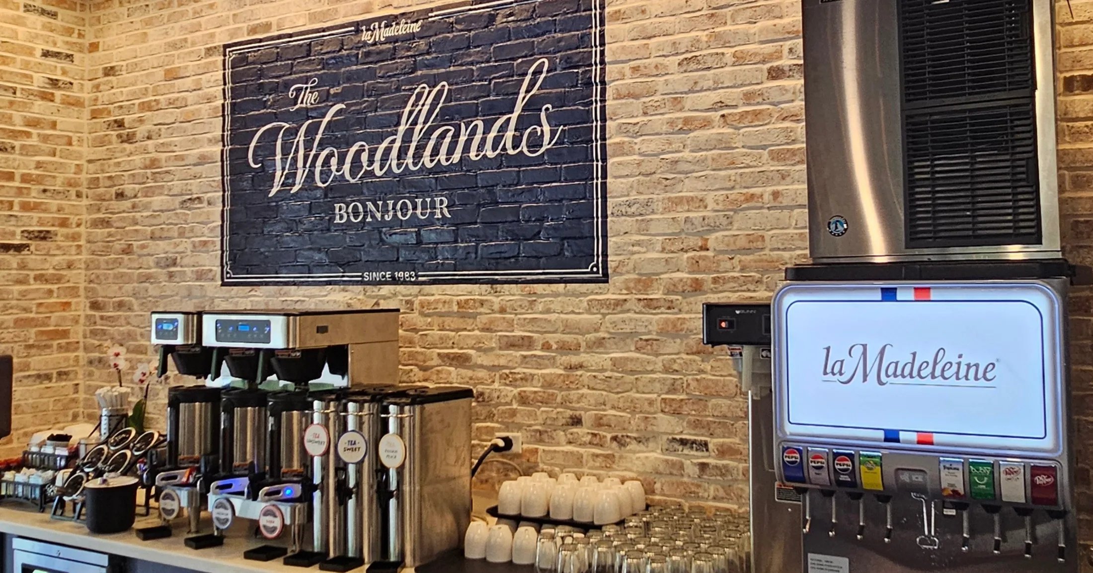 La Madeleine's beverage station with a custom The Woodlands Bonjour sign on an exposed brick wall