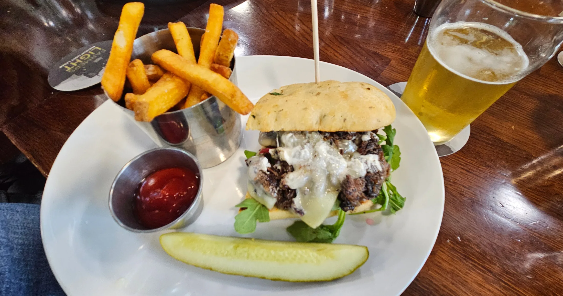 A shaved ribeye sandwich on focaccia bread with arugula and melted cheese, seasoned fries in a metal cup, pickle spear, and a pint of light beer at Farmers Brewing Co.