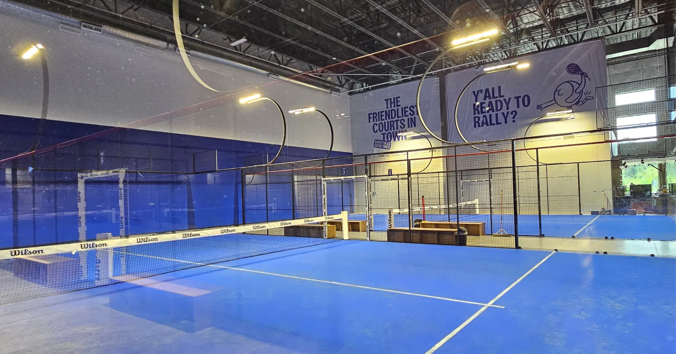 Wide interior view of multiple blue padel courts with Wilson nets, large overhead banners reading 'The Friendliest Courts in Town' and 'Y'all Ready to Rally?' visible above the courts