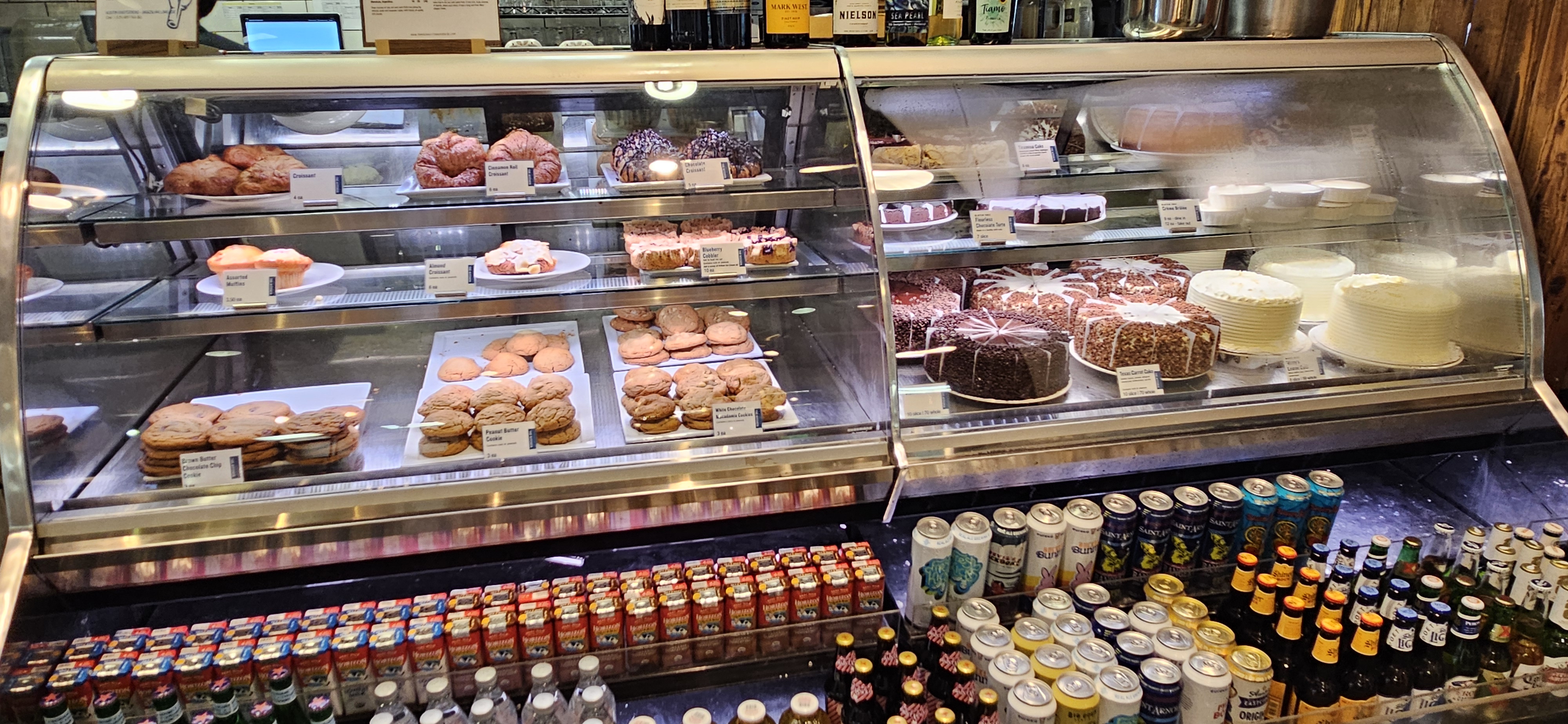Bakery display case at The Kitchen filled with croissants, cakes, muffins, peanut butter cookies, and whole layer cakes