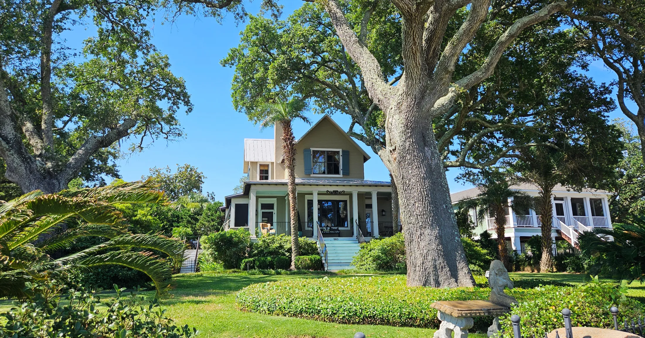 A charming two-story coastal cottage with blue shutters and a wrap-around porch, framed by large live oak trees and lush tropical landscaping in Gulfport, Mississippi