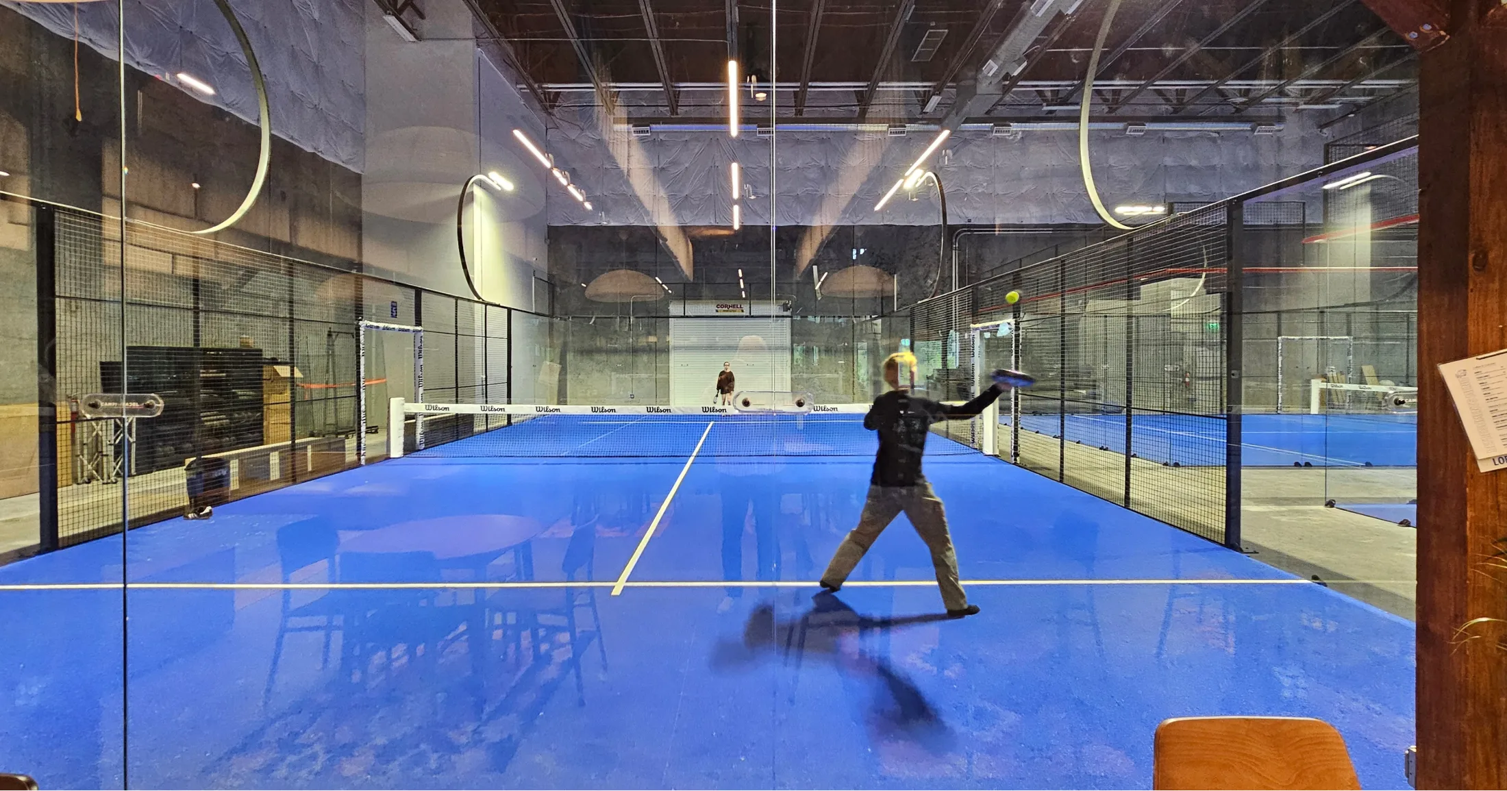 Player mid-swing on one of Lobb's five indoor padel courts, blue court surface and glass walls visible