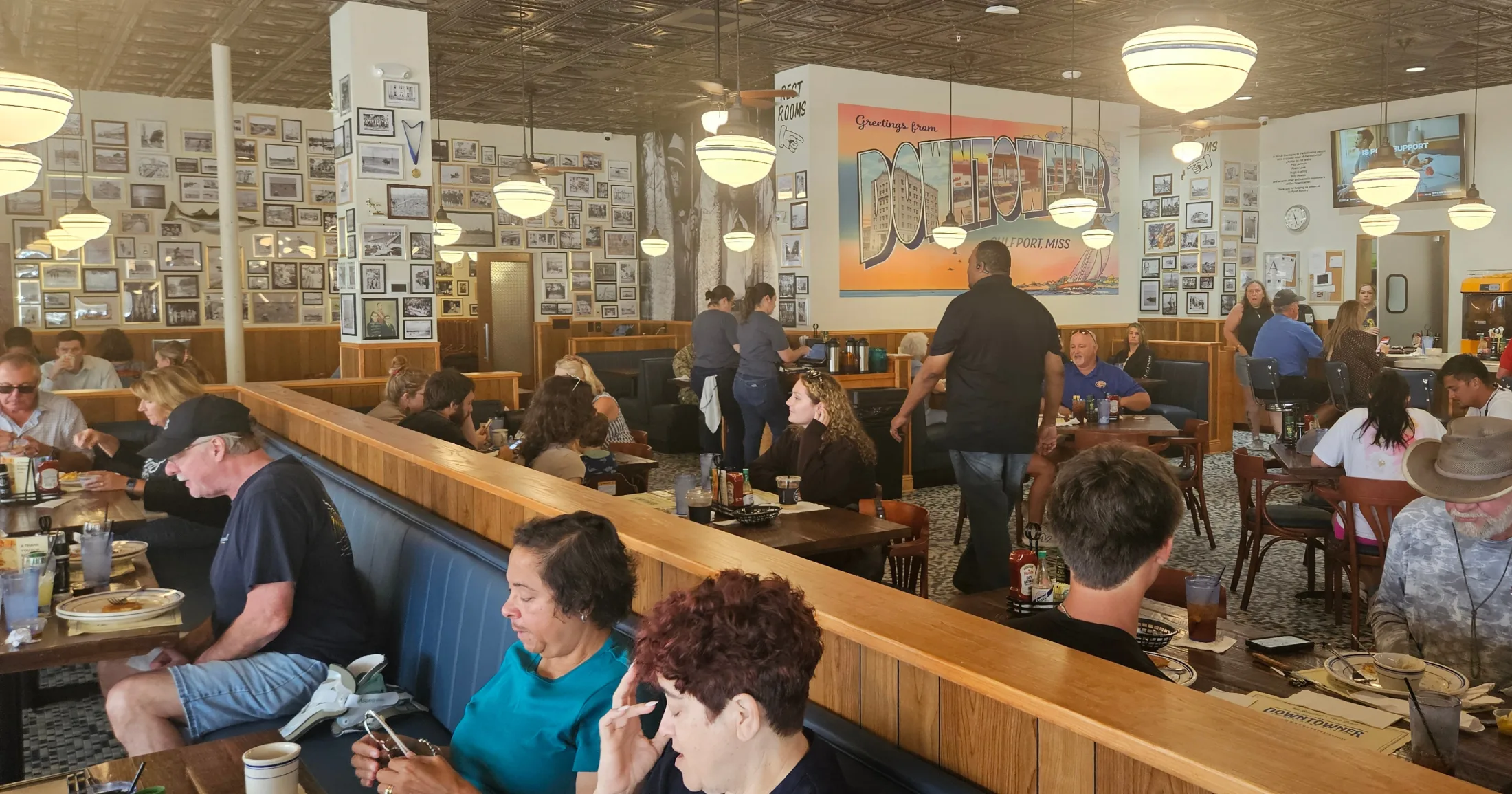 Packed dining room at The Downtowner with wood booths, vintage photo gallery walls, and a Greetings from Downtowner Gulfport mural