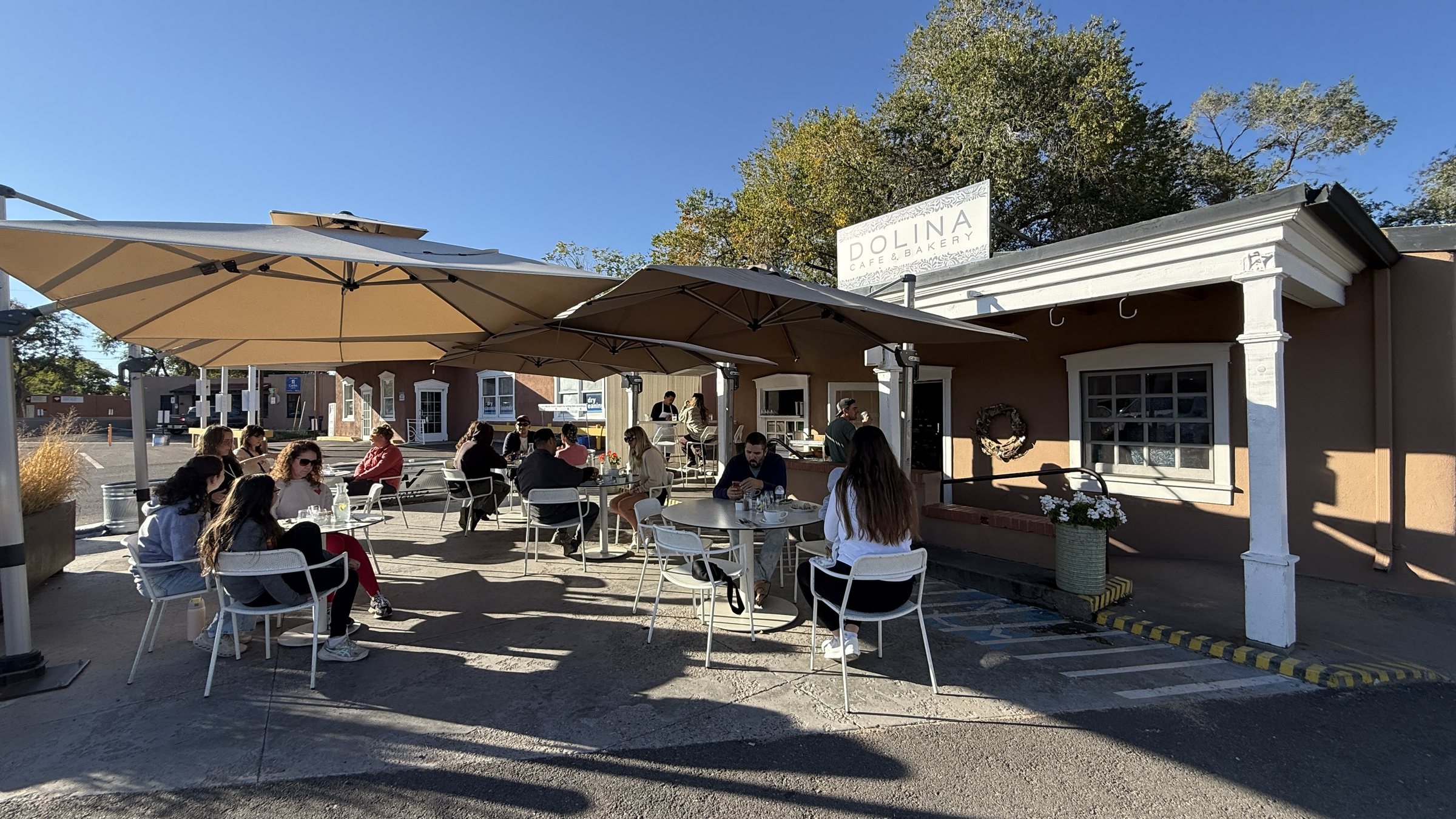 A sunny patio with white chairs and tan umbrellas beside a building with a sign reading Dolina Cafe & Bakery