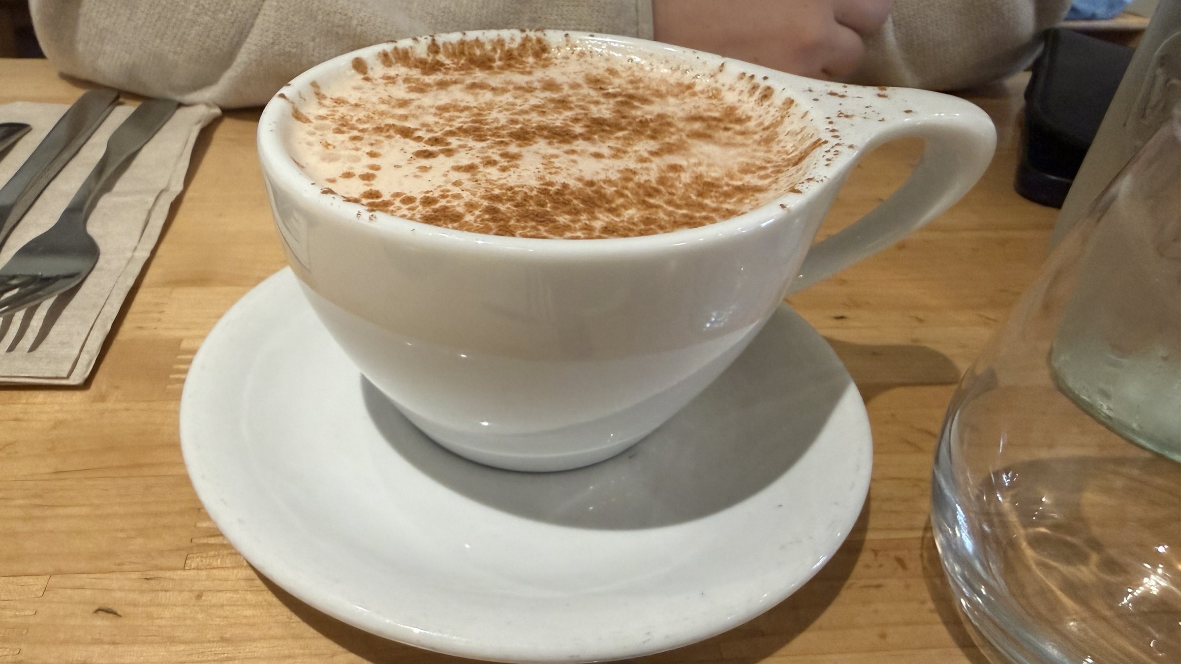 A hot drink in a big white cup and saucer dusted with cinnamon on a wooden table