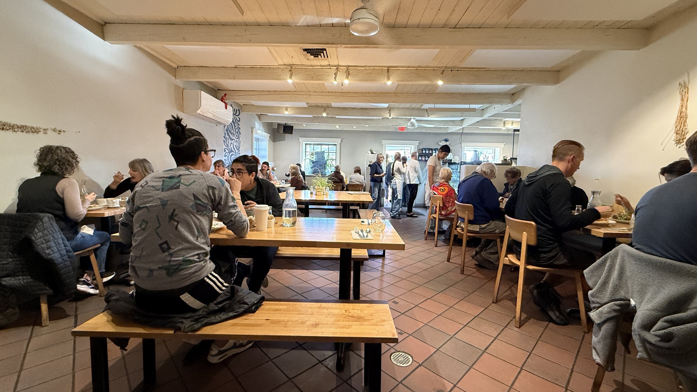 A busy dining room with white walls, white wood ceiling beams, a red tile floor and long wooden tables