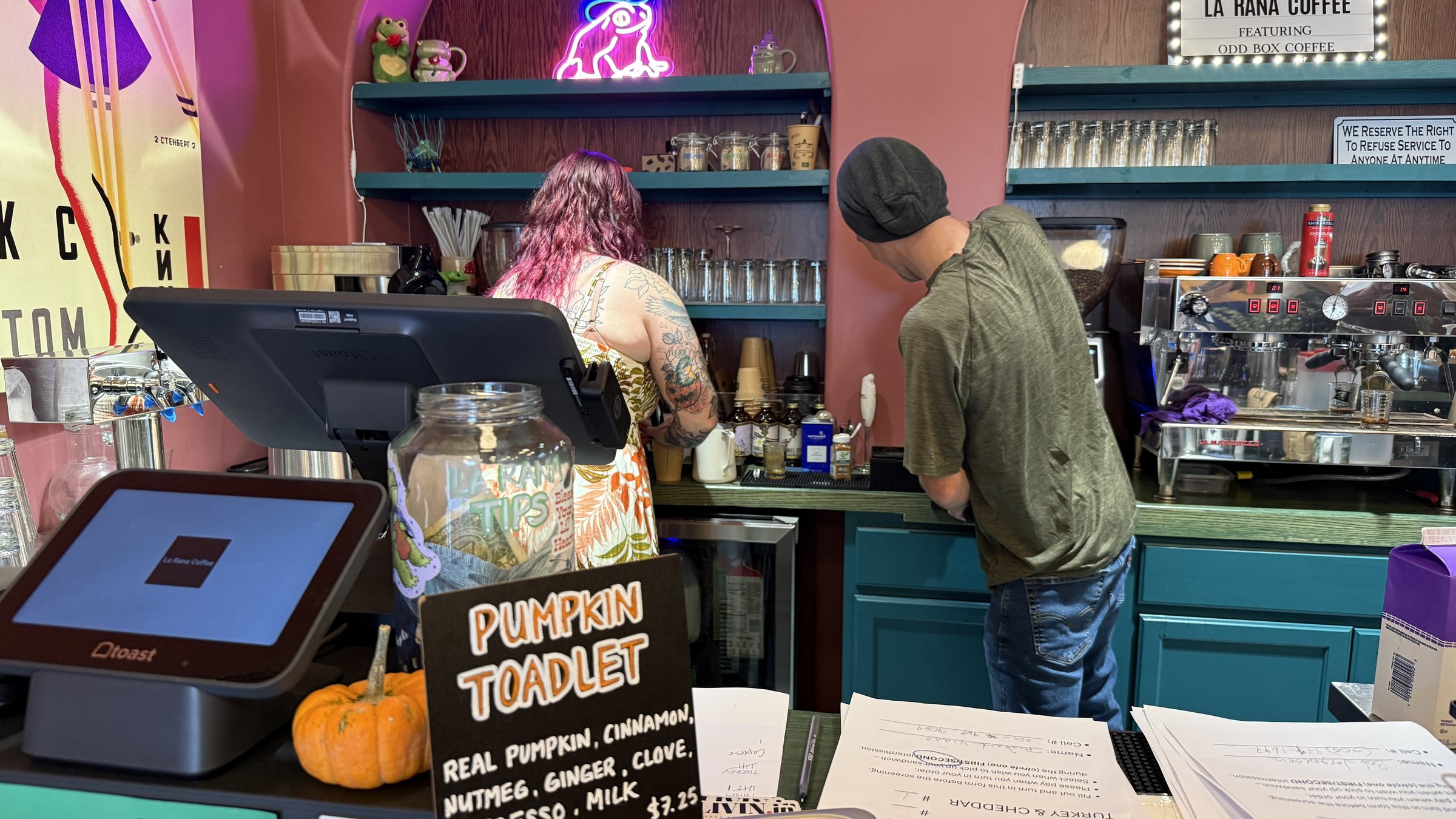 Two baristas at a coffee bar with teal shelves below a sign reading La Rana Coffee featuring Odd Box Coffee