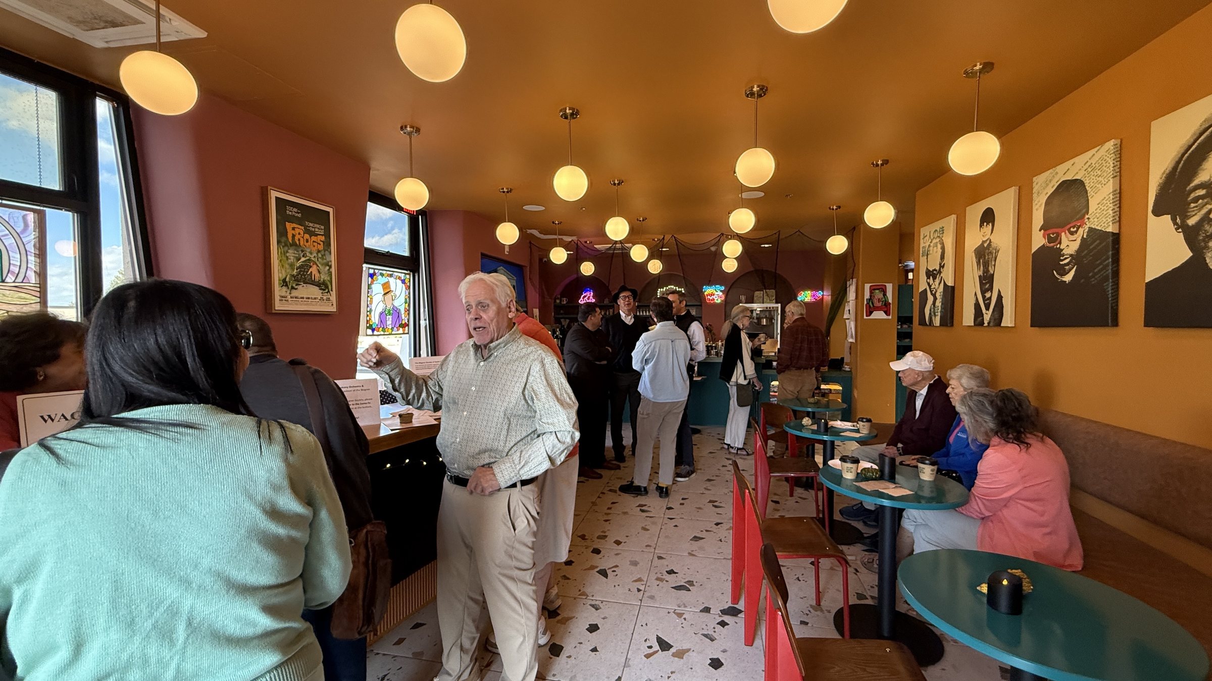A busy cafe lobby with globe pendant lights, a terrazzo floor, teal round tables, red chairs and black and white portraits