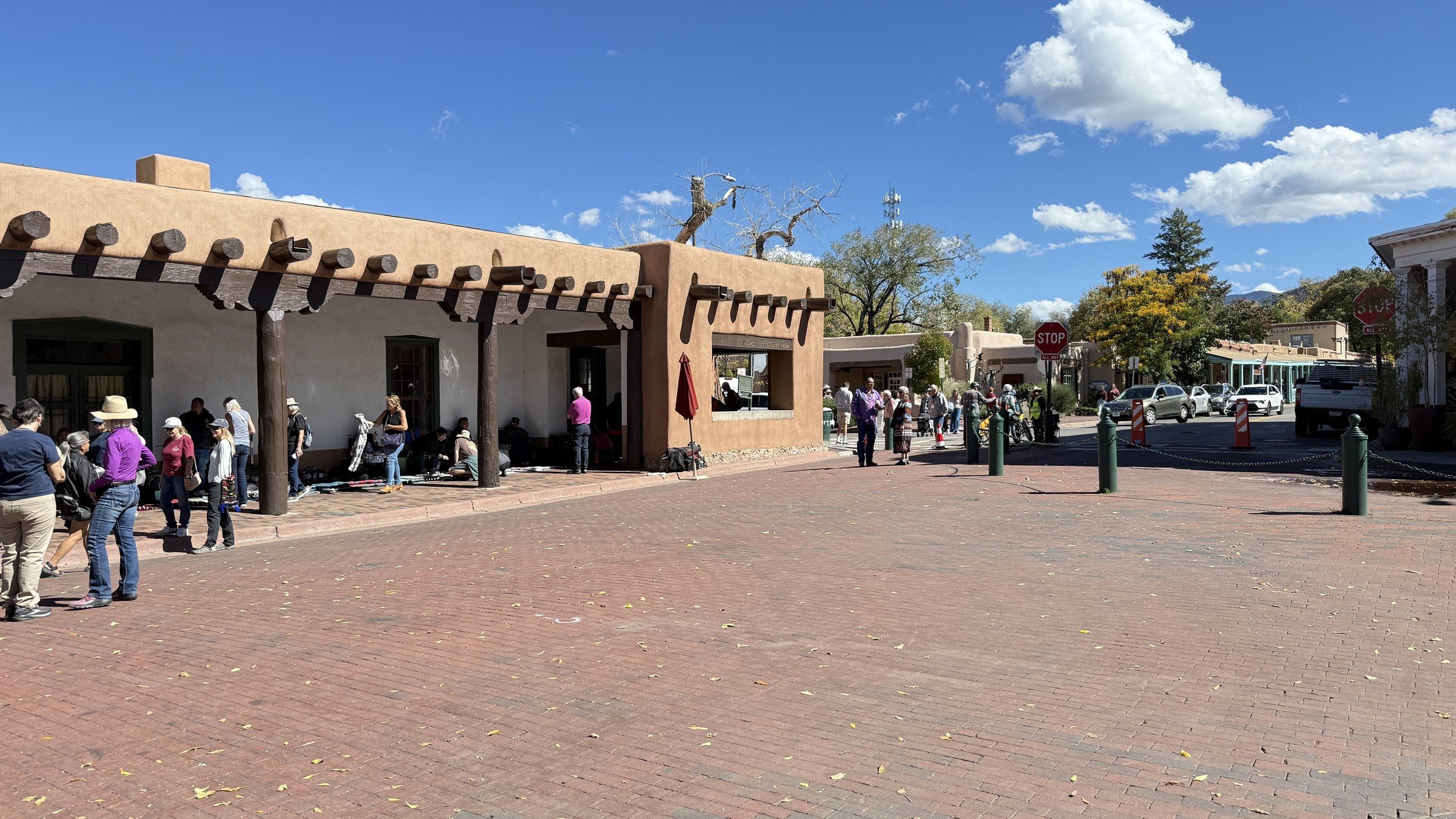 A brick plaza in front of a long adobe building with a wooden portal, with people walking and standing under a blue sky