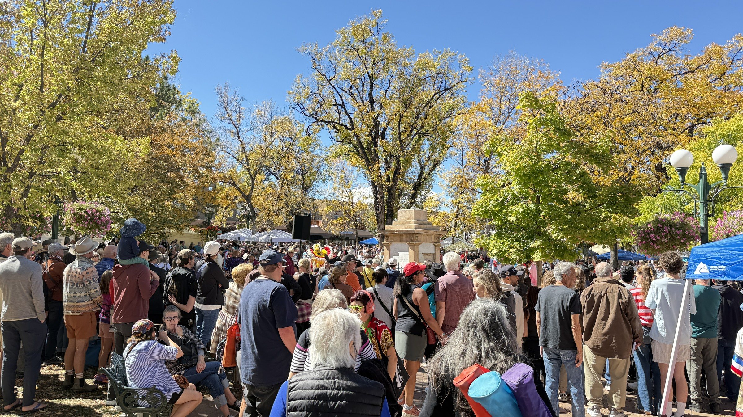 A crowd gathered under tall trees with yellow and green leaves on the Santa Fe Plaza, with a white tent and lamp posts
