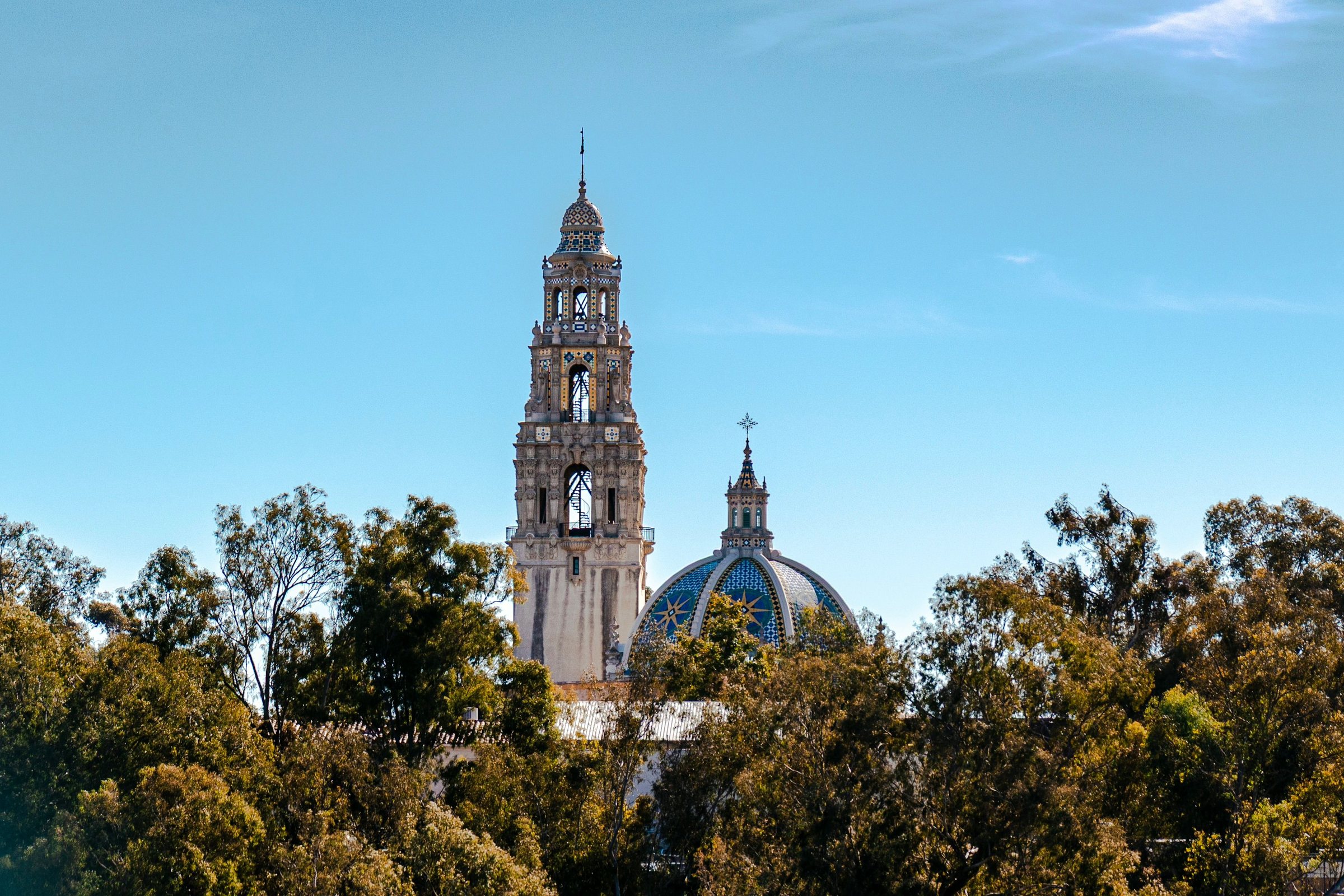 A tall ornate tower with several tiers and a pointed top rising above green treetops next to a blue and gold tiled dome, under a blue sky