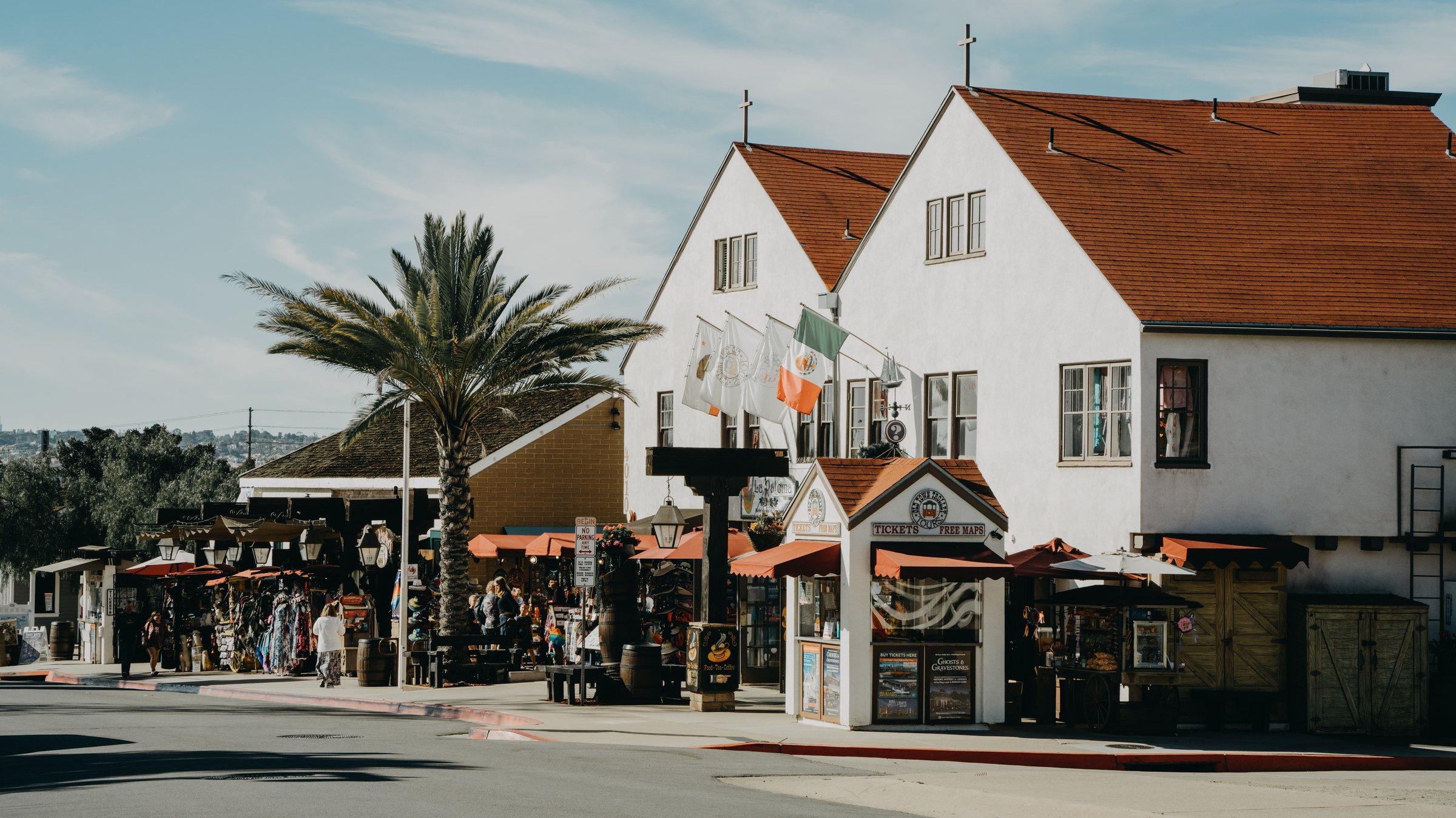 A sunny street corner with a white two-story building with a red-brown roof, shop awnings, a palm tree and people walking on the sidewalk