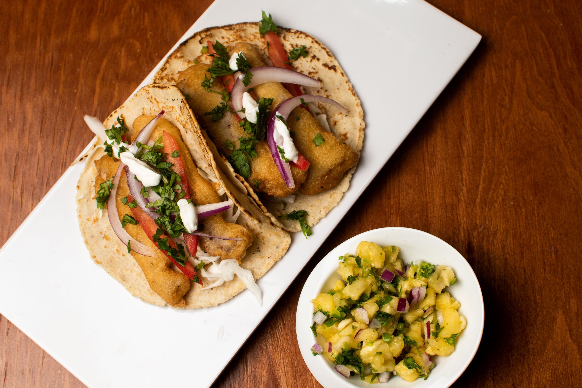 Two fish tacos on a white plate served with a small bowl of pineapple salsa on a wooden table